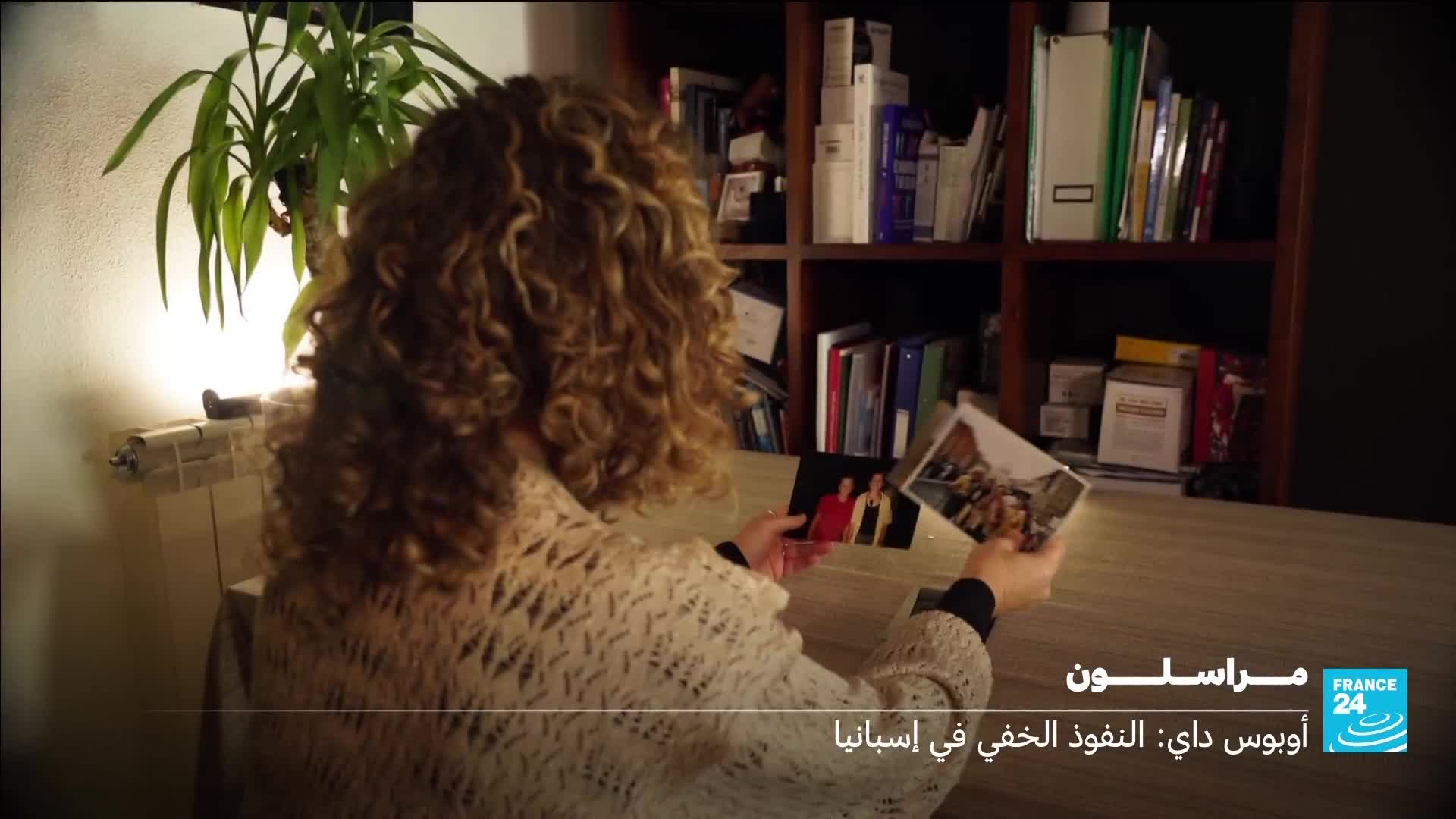 A woman with curly hair holds two photographs, one showing two people in red and yellow, the other a group. The France 24 logo appears in the lower right corner. A woman with curly hair holds two photographs, one showing two people in red and yellow, the other a group. The France 24 logo appears in the lower right corner.