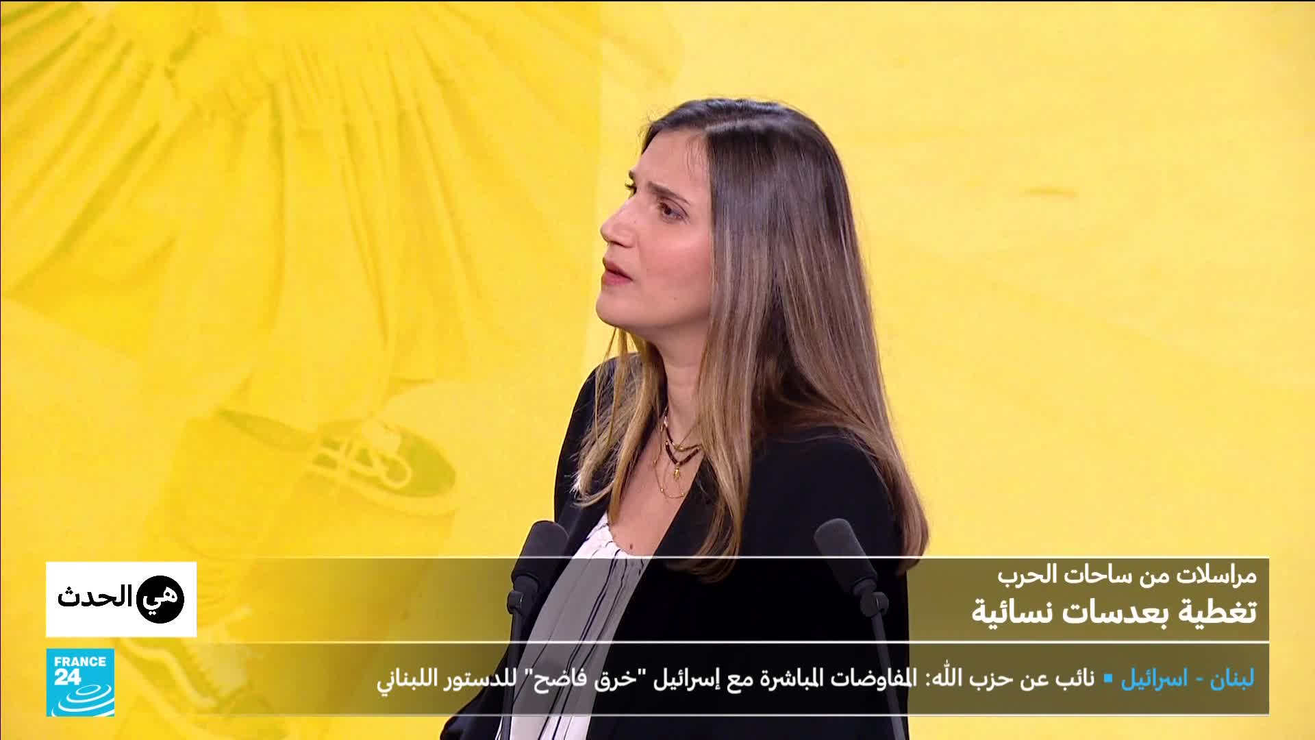 A woman with long brown hair speaks into a microphone, her expression serious. Behind her, a bright yellow backdrop features a subtle pattern of what looks like fabric folds.