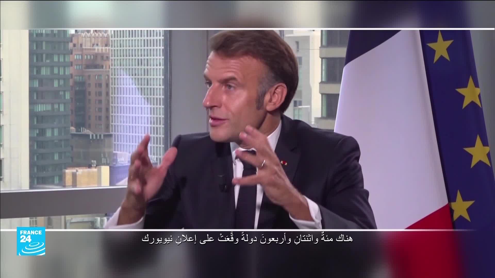 The French president gestures with both hands as he speaks, a French flag with European stars behind him. He's in a room with a large window overlooking city buildings.