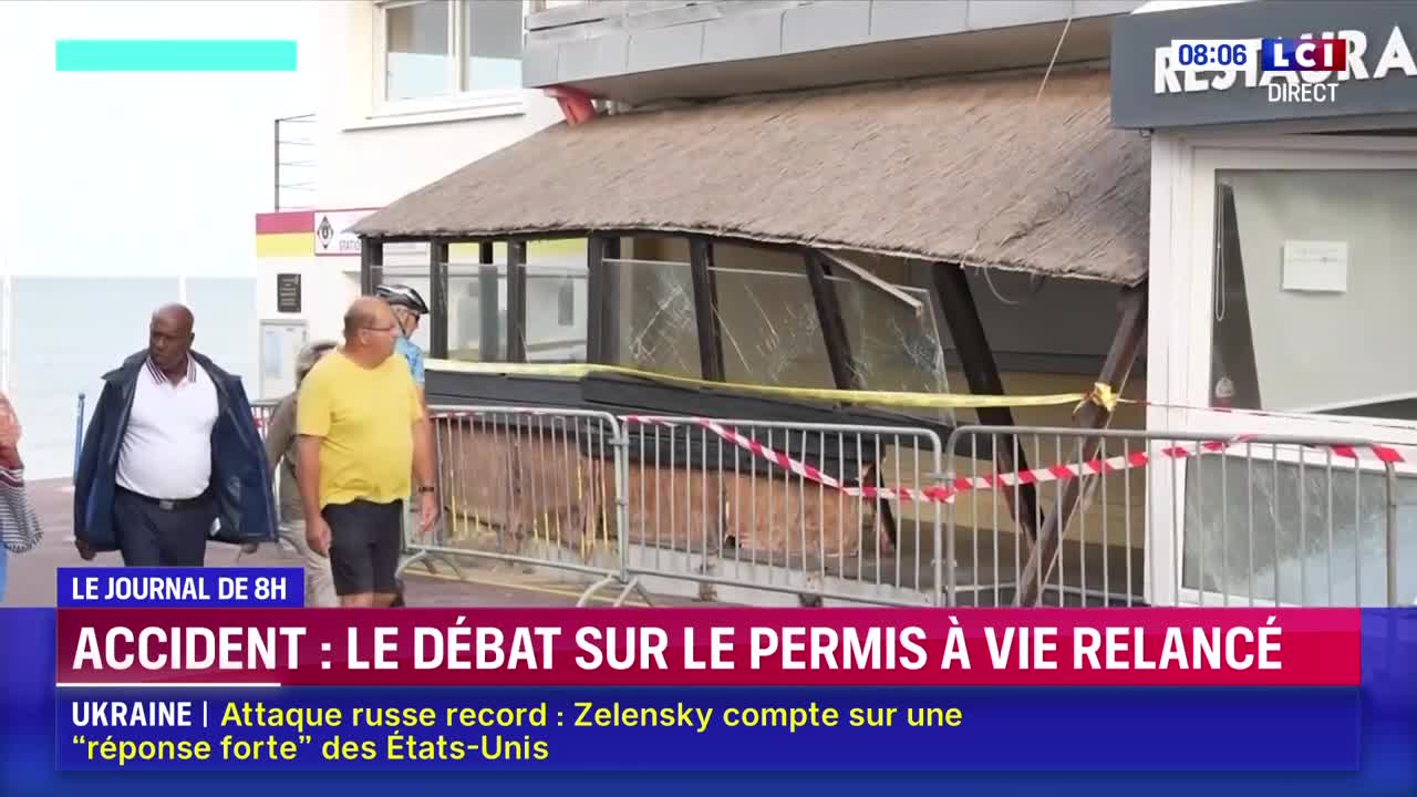 A restaurant's window is shattered, with red and white tape cordoning off the damage. People stroll past the scene as LCI's coverage of the accident airs on the screen.

