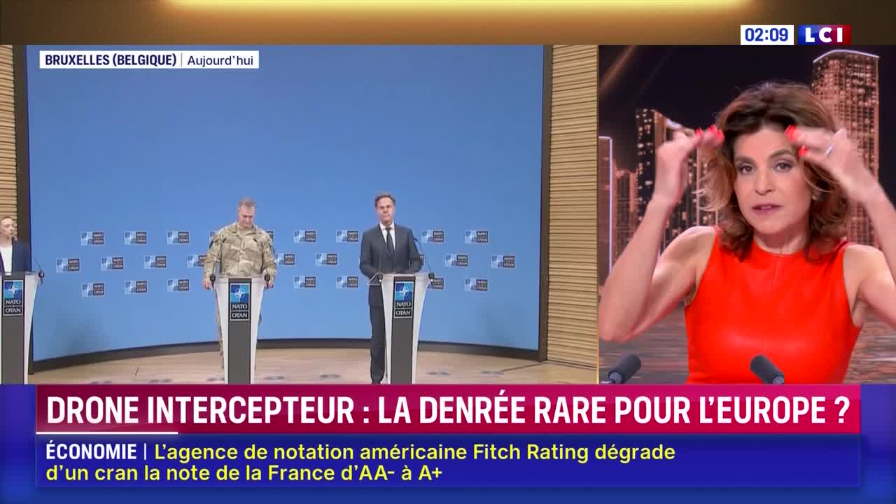 A woman in a bright red dress gestures with both hands as she speaks. Behind her, a news segment on LCI shows a panel in Brussels discussing drones.
