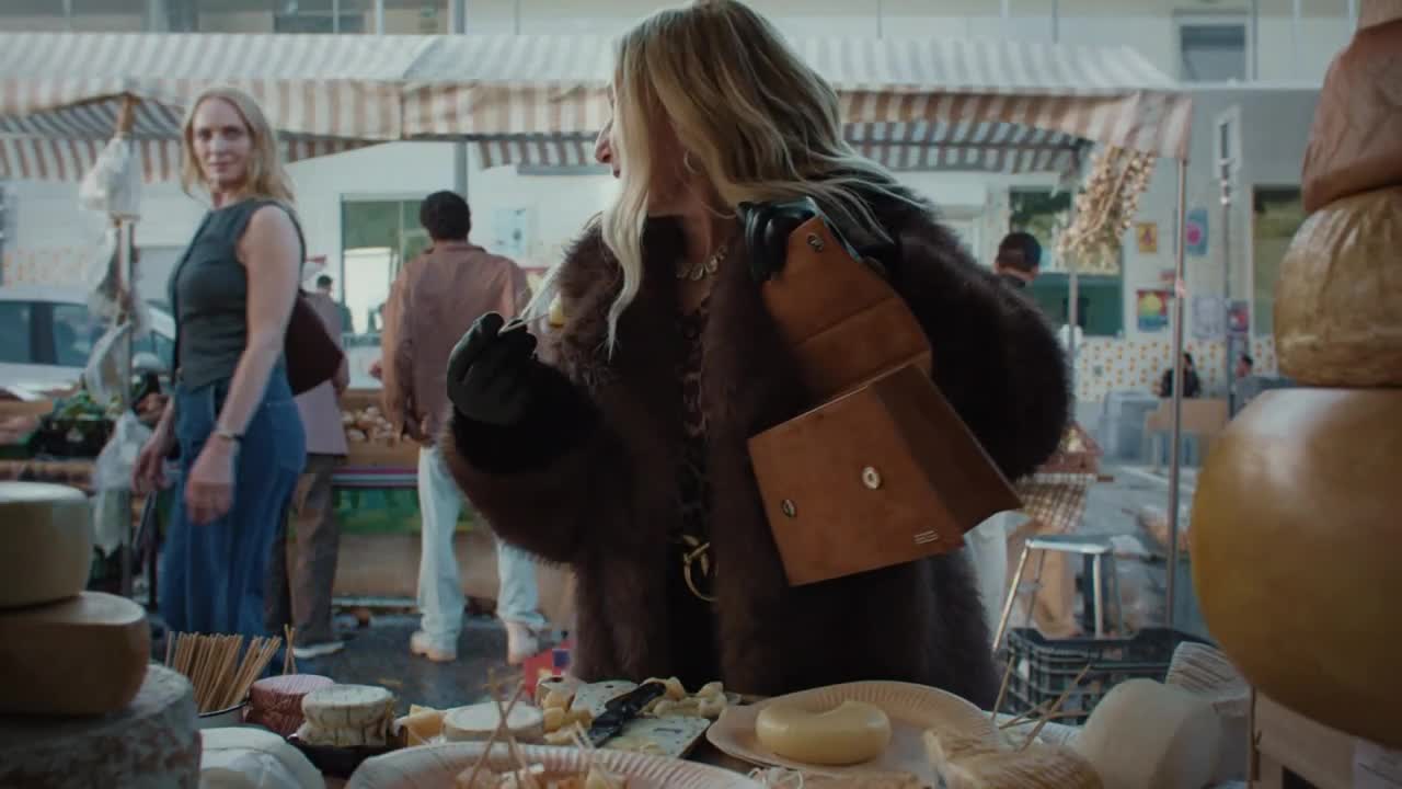 A woman in a fur coat, leather gloves, and a purse examines cheese at a market stall. Another woman in a tank top and jeans looks on, standing nearby.
