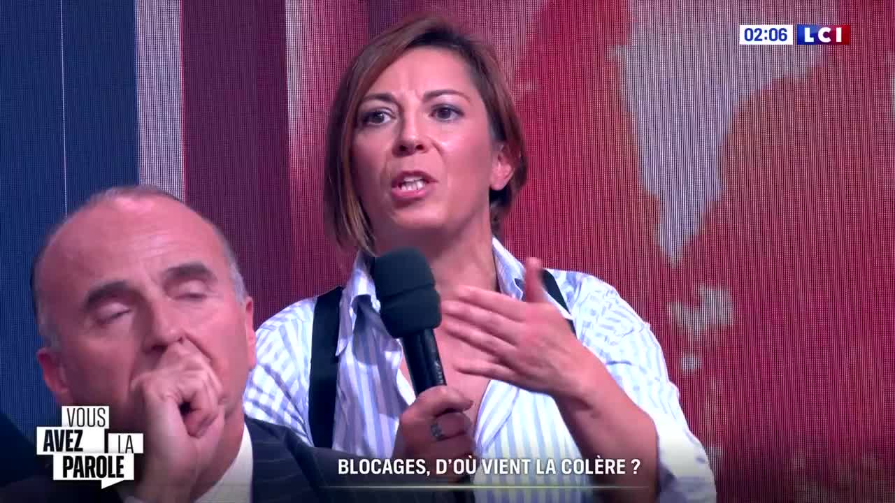 A woman with short brown hair gestures emphatically while speaking into a microphone. Beside her, a man in a suit appears to be listening intently, hand near his face.
