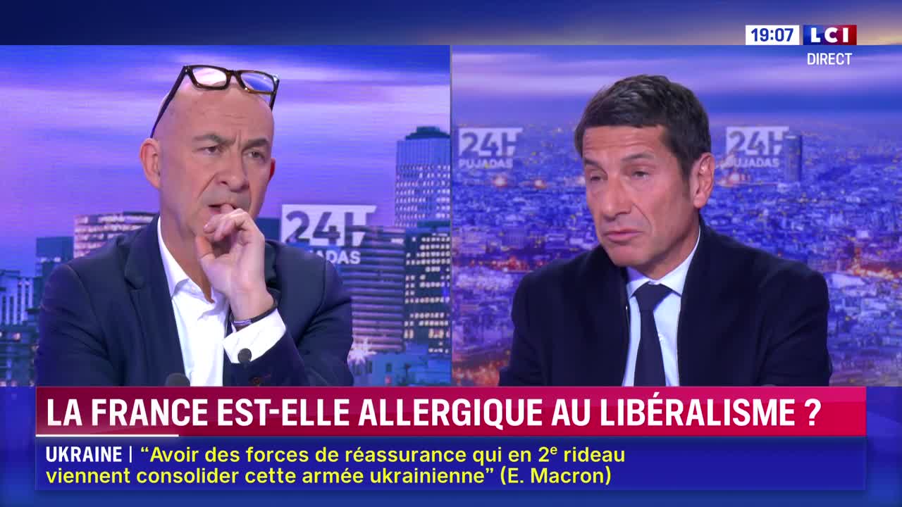 Two men in suits sit at a desk on the LCI set, facing the camera. The man on the left has his glasses perched on his head, and he's touching his chin thoughtfully.
