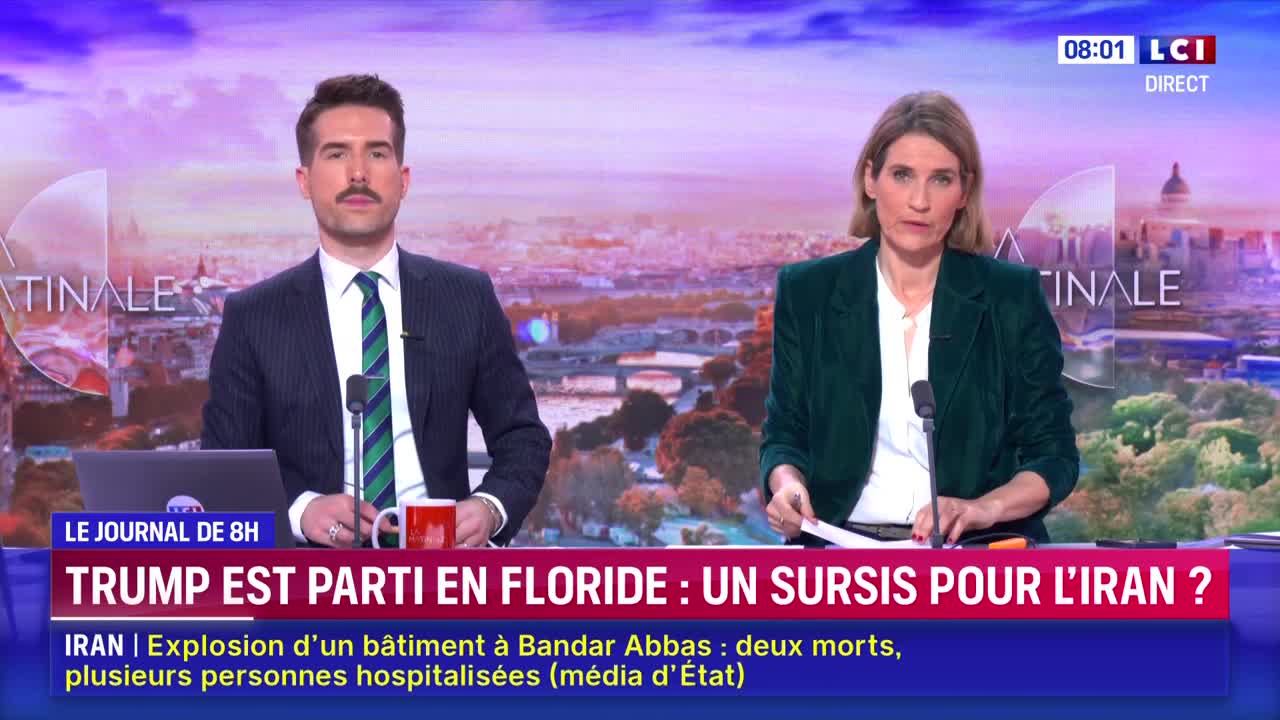 The LCI news anchors are seated at their desk, with a city skyline behind them. The male anchor, sporting a mustache, gestures with his hands while the female anchor looks down at her papers.