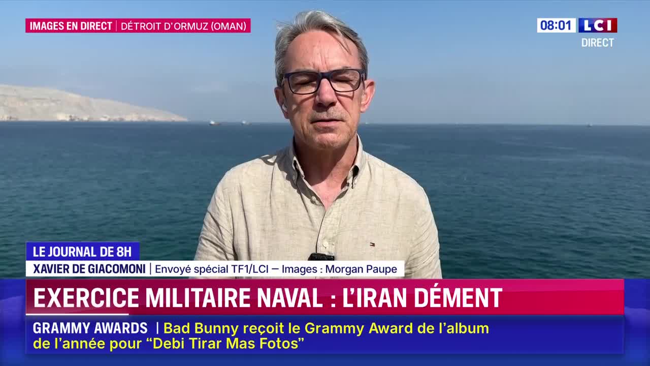 Several ships are scattered across the calm, blue water of the Strait of Hormuz. A man in a tan shirt stands before the sea, reporting live for LCI.