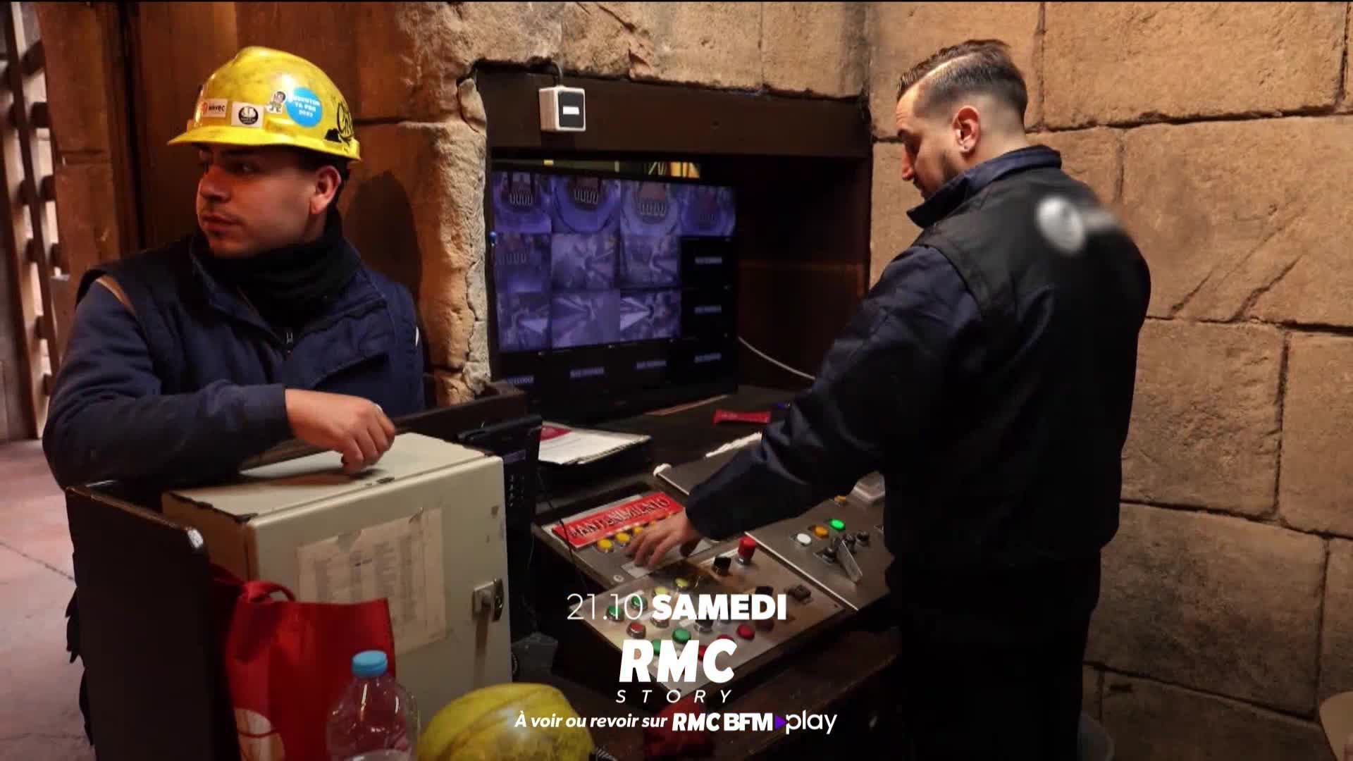 A man in a dark uniform is focused on a control panel, while another, wearing a yellow hard hat, looks off to the side. The scene, likely from a French RMC Story broadcast, is set inside a stone-walled room.
