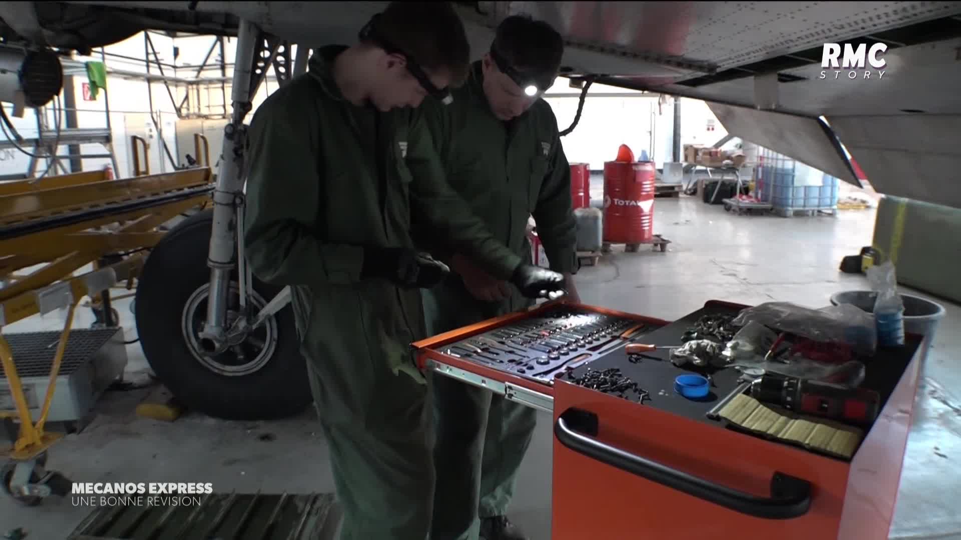 Two mechanics in green coveralls, illuminated by headlamps, pore over a toolbox filled with small parts, likely under the belly of an aircraft. The scene, captured by RMC Story in France, seems to be part of a maintenance check.
