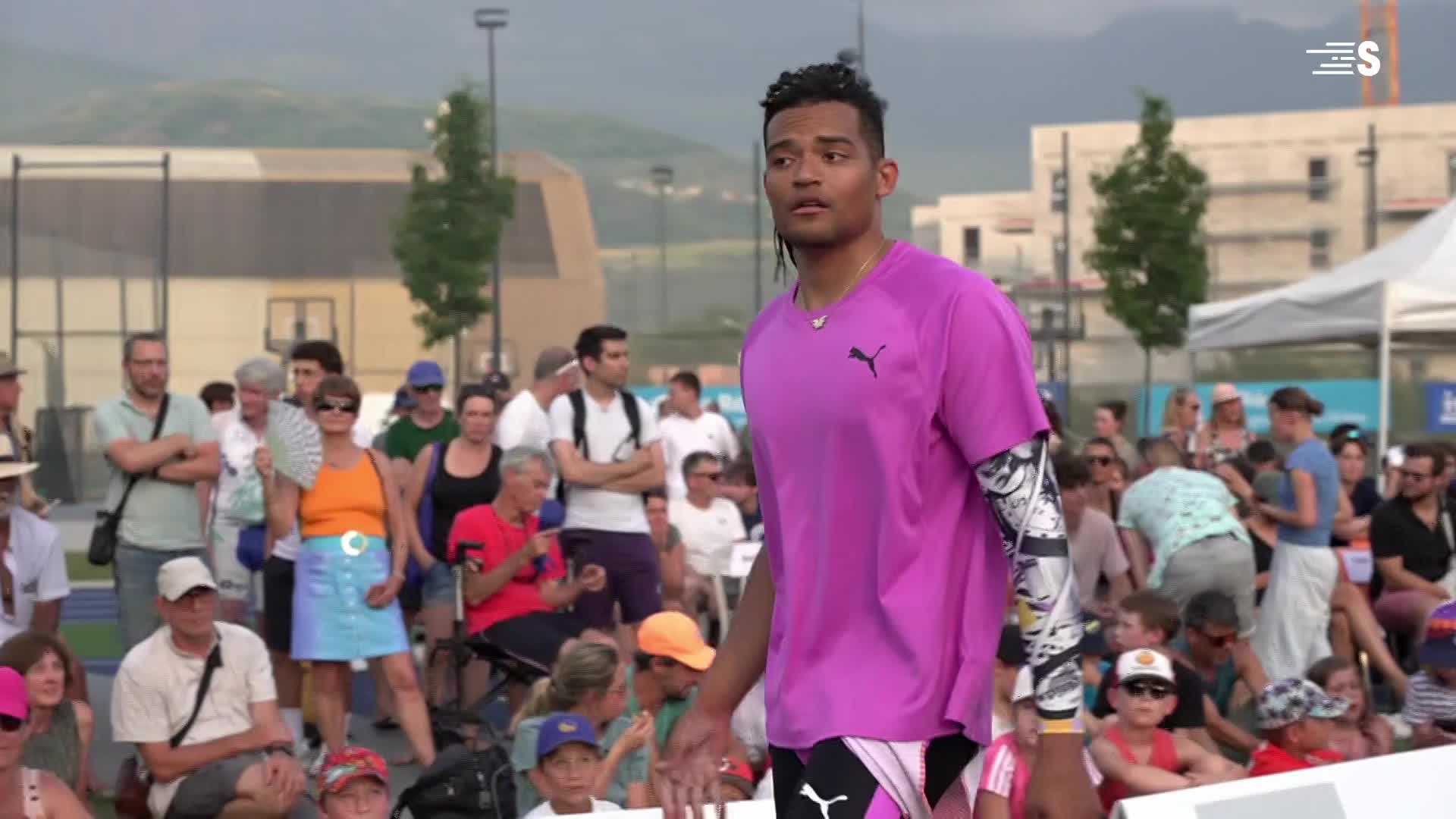 A track athlete, wearing a bright purple Puma shirt, surveys the crowd after a competition. Spectators, some with fans, watch from the stands under the summer sun in France.
