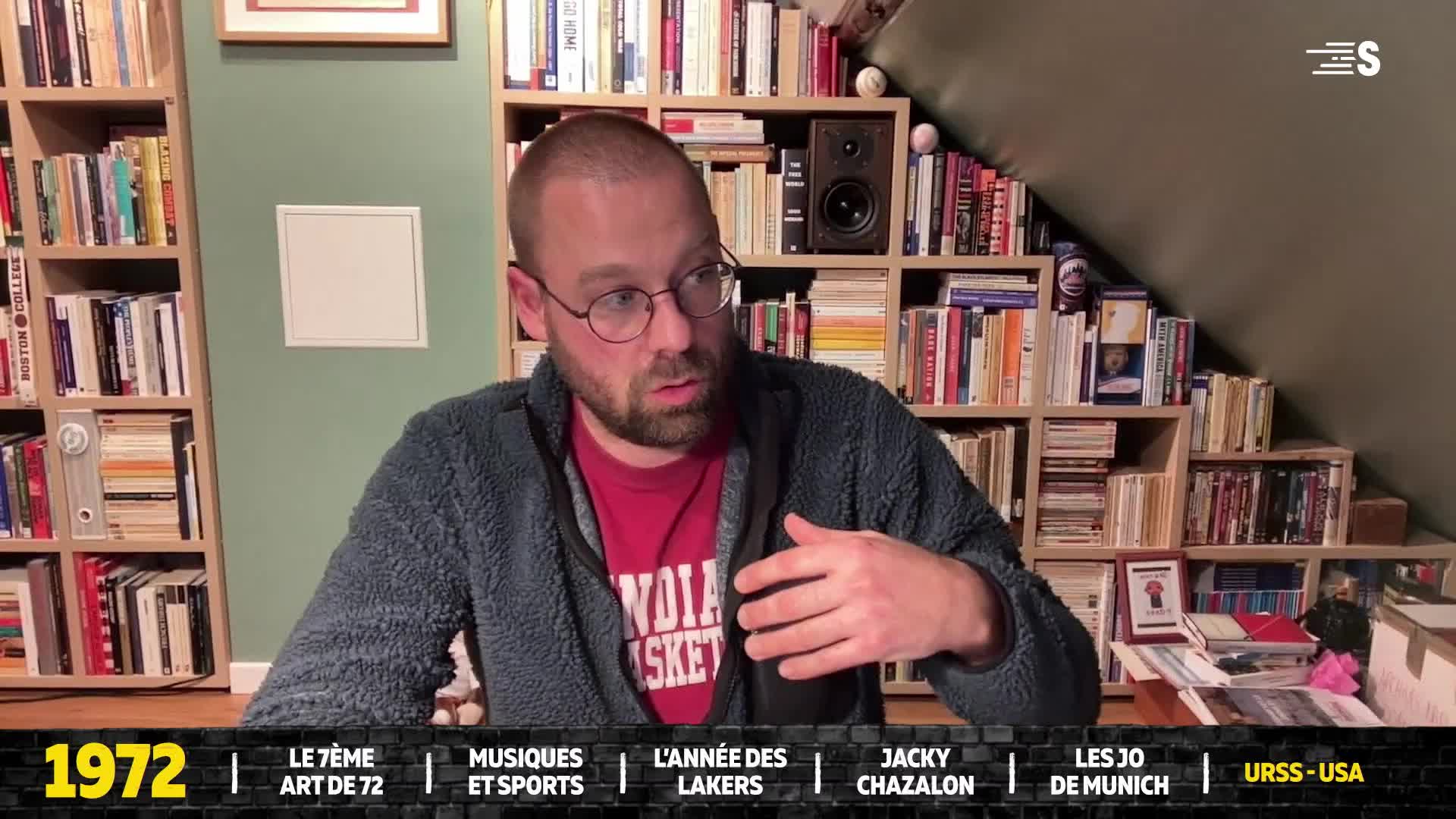A man in glasses and a red shirt is speaking, his hands gesturing as he sits in front of a bookshelf.  Below, a timeline from Sport en France highlights various topics from 1972.
