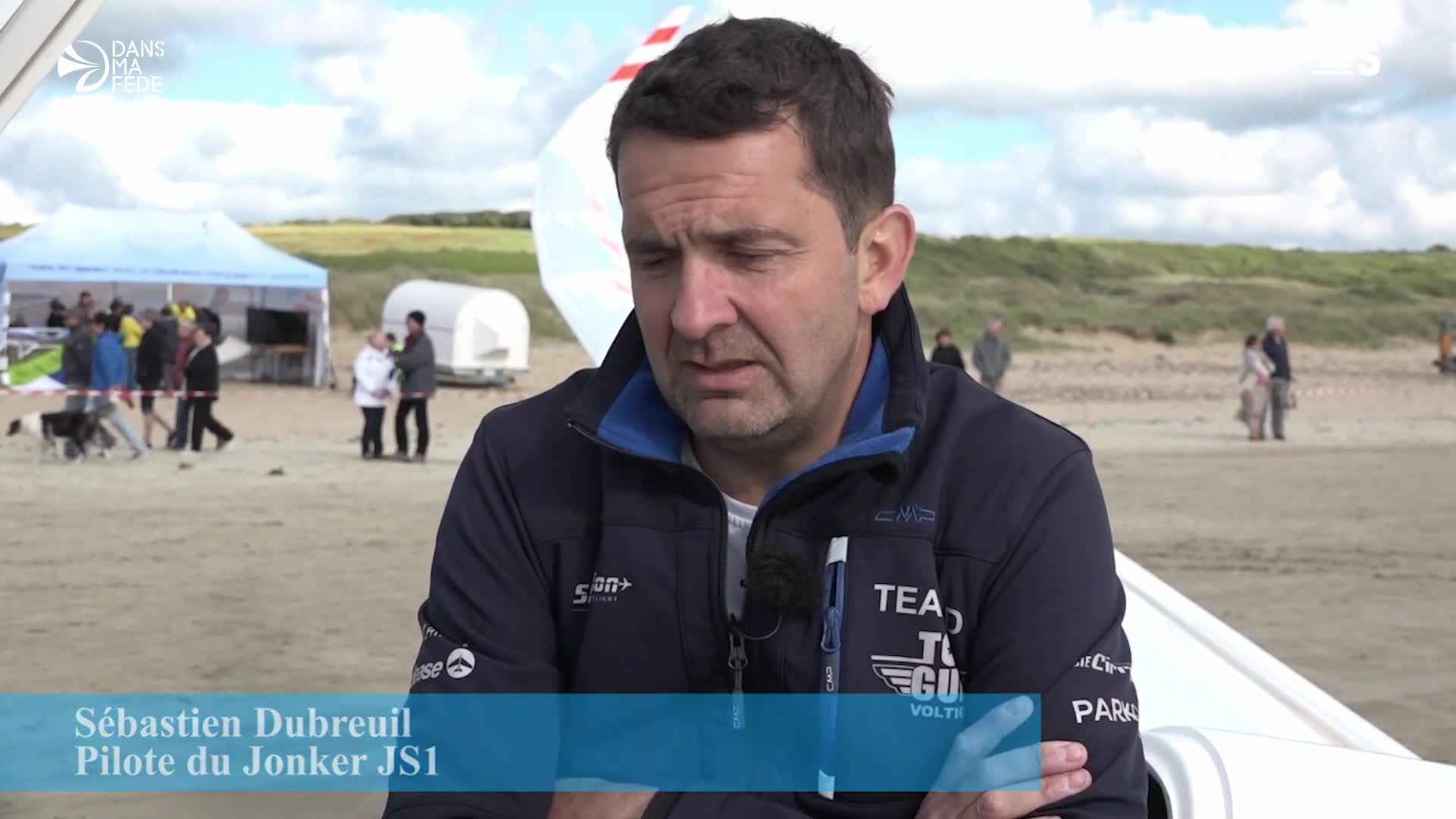 Sébastien Dubreuil, the Jonker JS1 pilot, speaks to the camera on a sandy beach, likely for Sport en France. Behind him, people mill about near a tent and a glider's wing.
