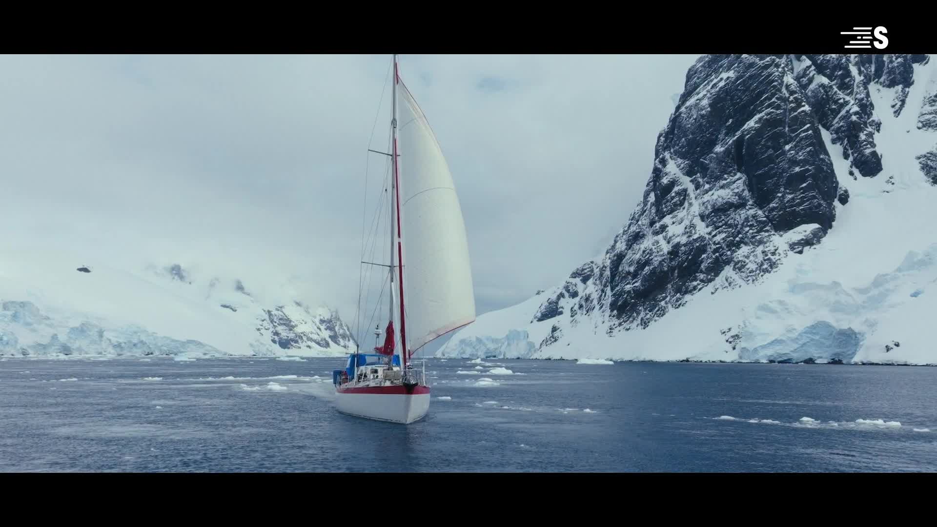 A sailboat with a white sail cuts through the icy water, heading toward a snow-covered mountain. The red trim on the boat pops against the stark, cold landscape.
