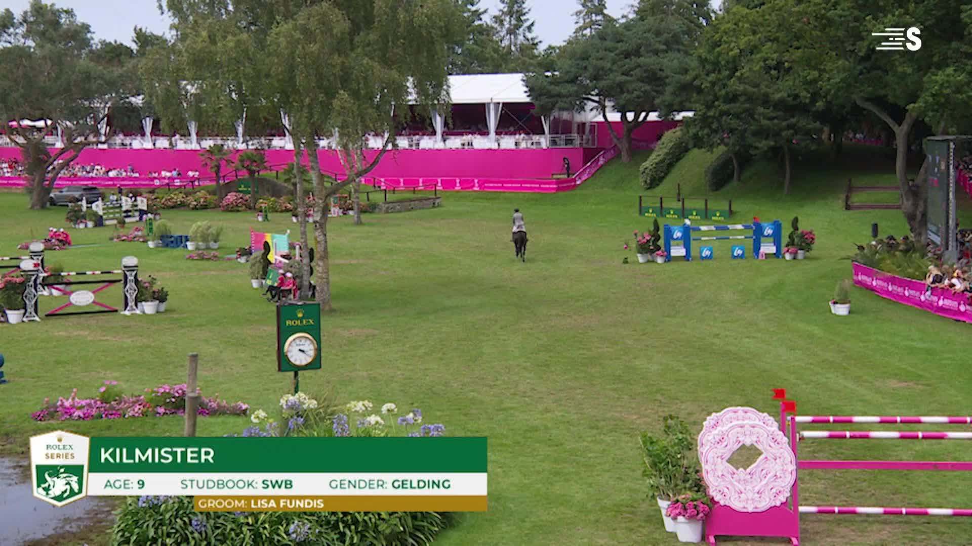 A horse and rider are approaching a set of blue jumps, navigating the grassy course. The event is taking place outdoors, with spectators and colorful decorations lining the perimeter.

