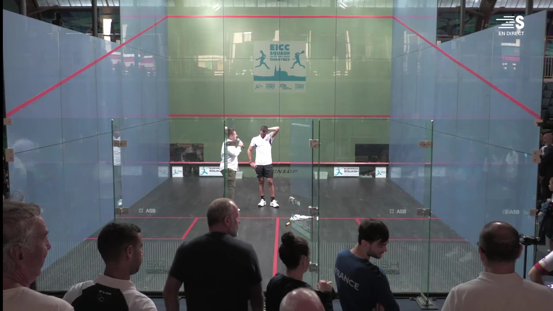 The squash players stand on the court, one with his hand on his head, the other talking. Spectators watch the match in France, broadcast live on Sport en France.
