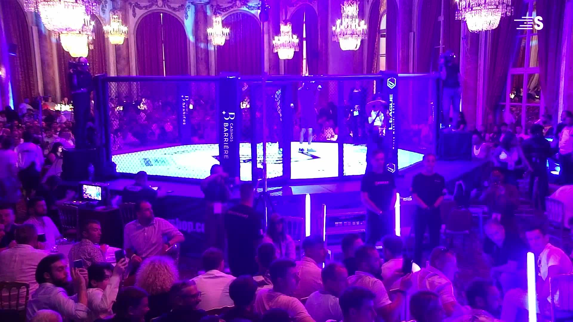 Inside the cage, a fighter stands, lit by purple spotlights. Chandeliers hang above the crowd in the opulent hall, where spectators watch the event in France.
