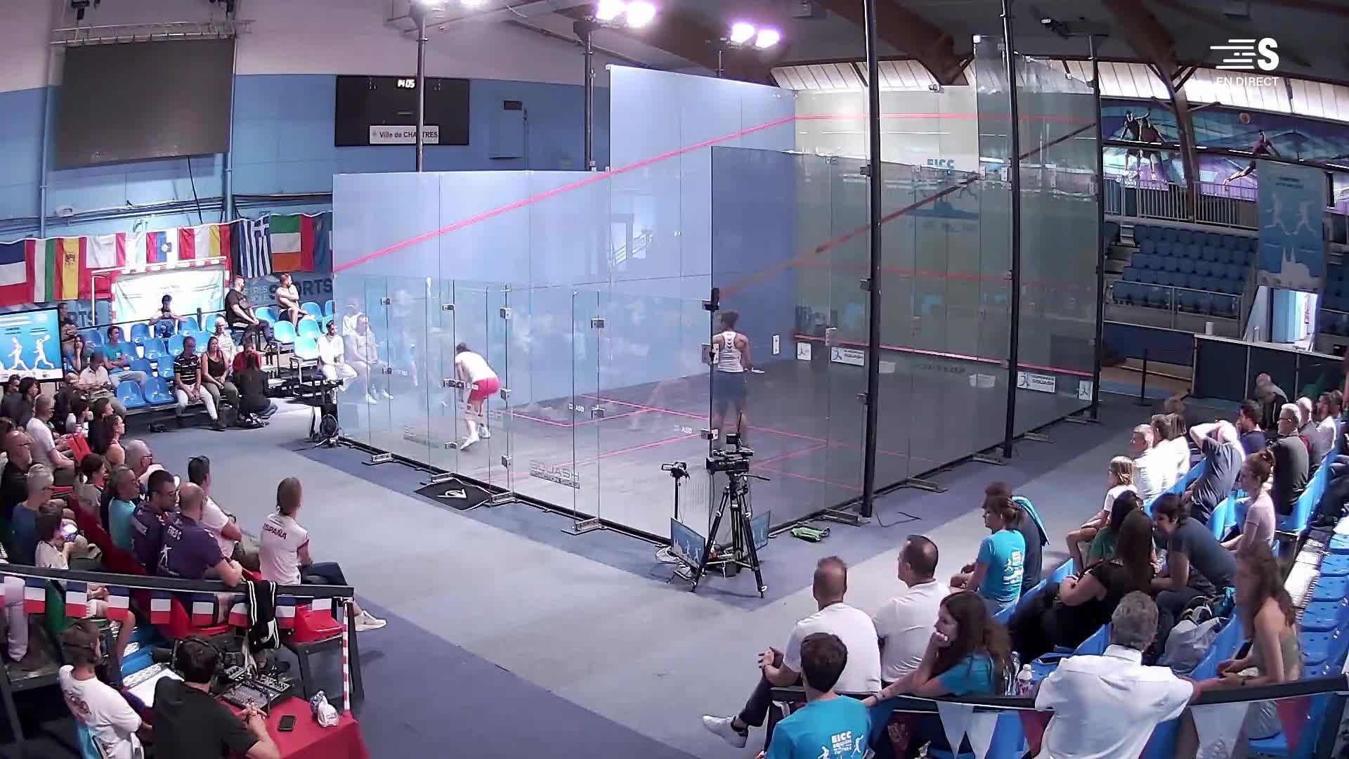 Two squash players, clad in athletic gear, are locked in a rally inside the glass court. The crowd watches intently as one player, in pink shorts, returns the ball. 
