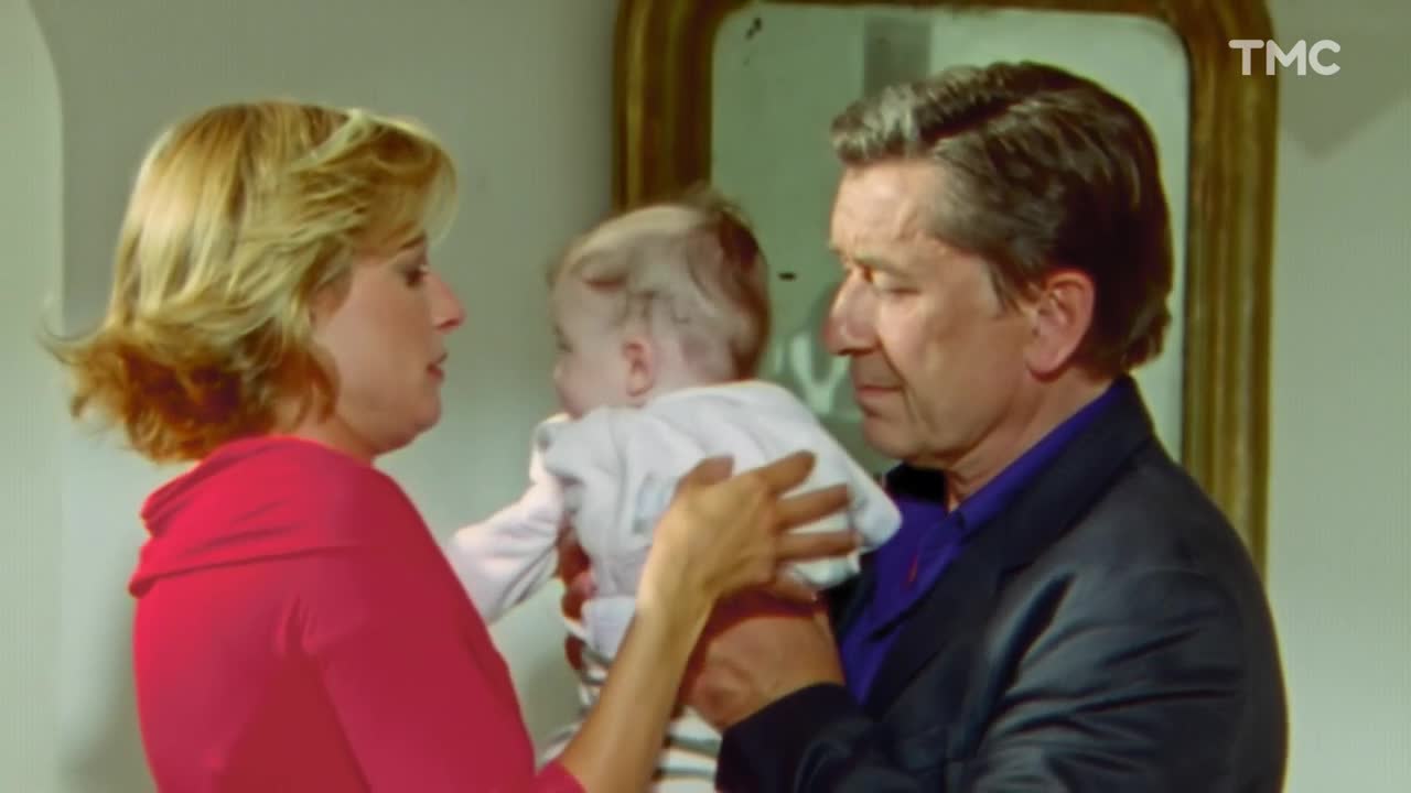 A woman in a red top and a man in a suit are holding a baby between them. The baby, in a white onesie, is facing away from the camera.
