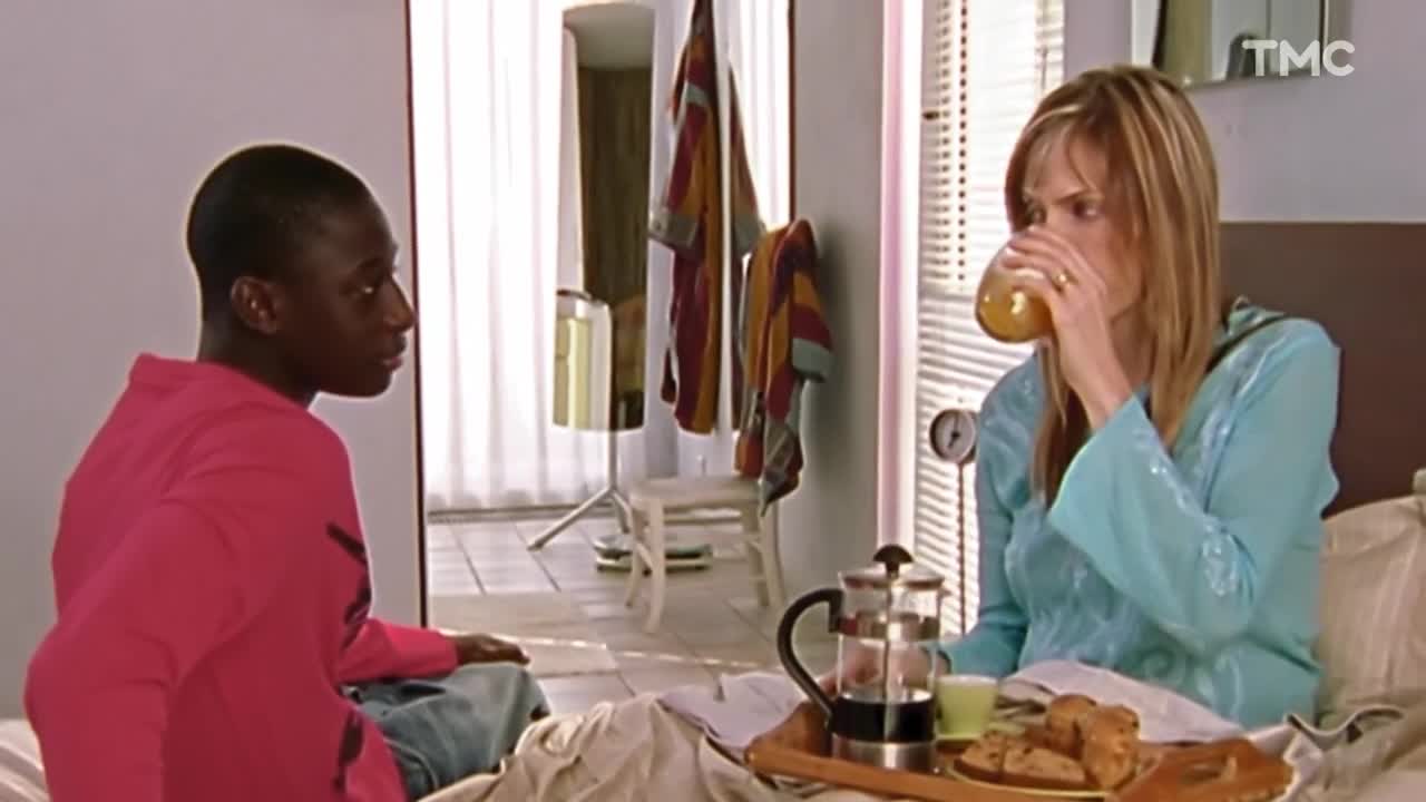 A young man in a pink shirt sits on the bed, looking towards a woman. She takes a drink from a glass, a breakfast tray in front of her.
