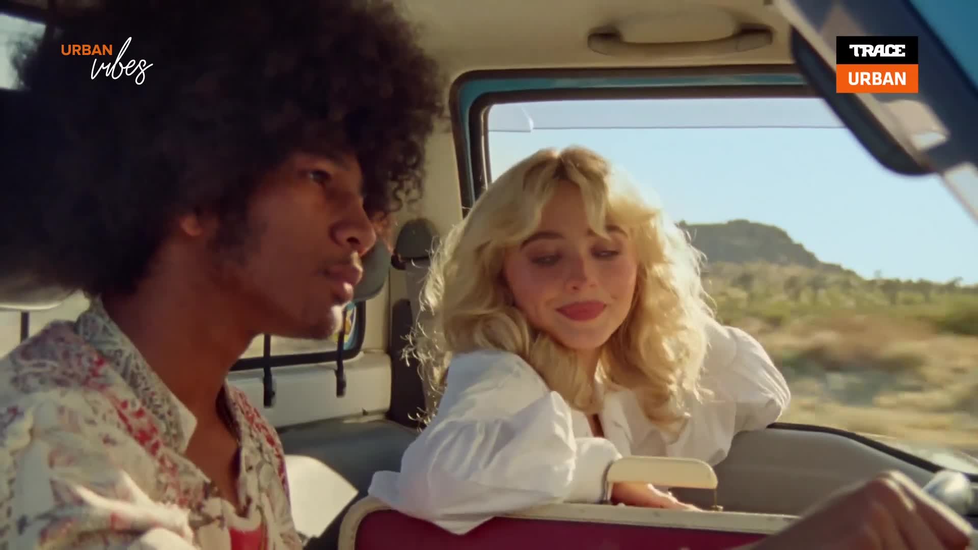 The woman leans out the car window, smiling at something off-camera. Beside her, the man with the big hair looks in the same direction, his expression thoughtful.
