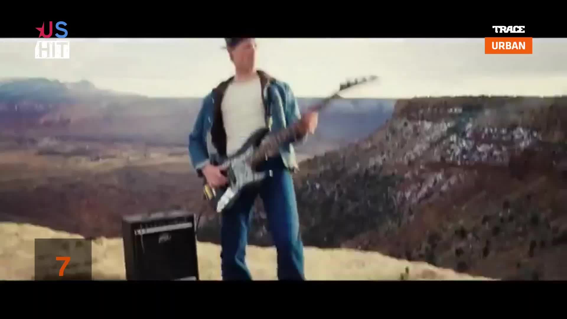 A man in a denim jacket strums a guitar, standing on a plateau. Behind him, a vast, arid landscape stretches towards the horizon, and the Trace Urban logo sits in the corner.
