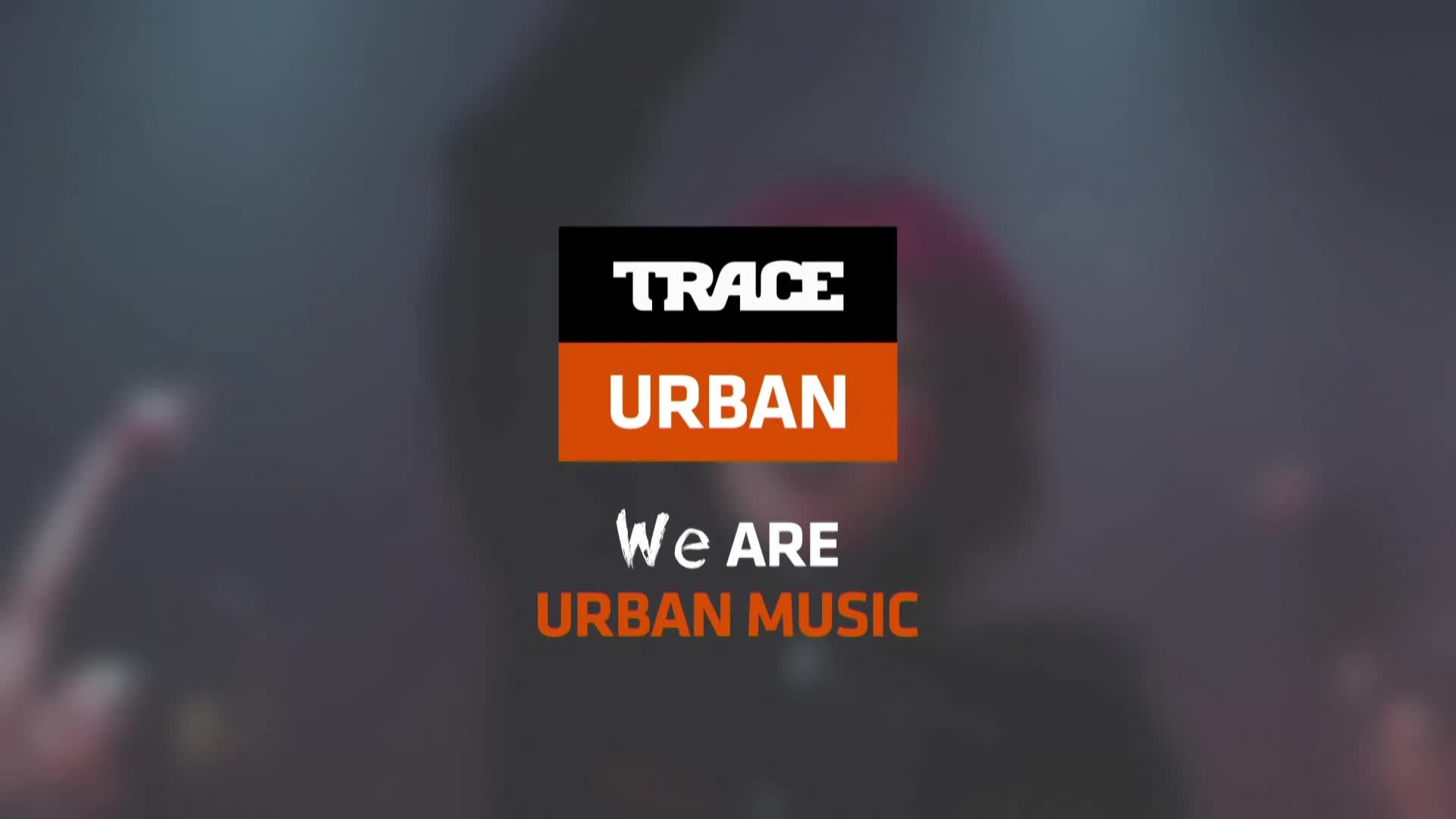 The Trace Urban logo sits front and center, with a dark background. A crowd is blurred in the background, and someone's arm is raised in the air.
