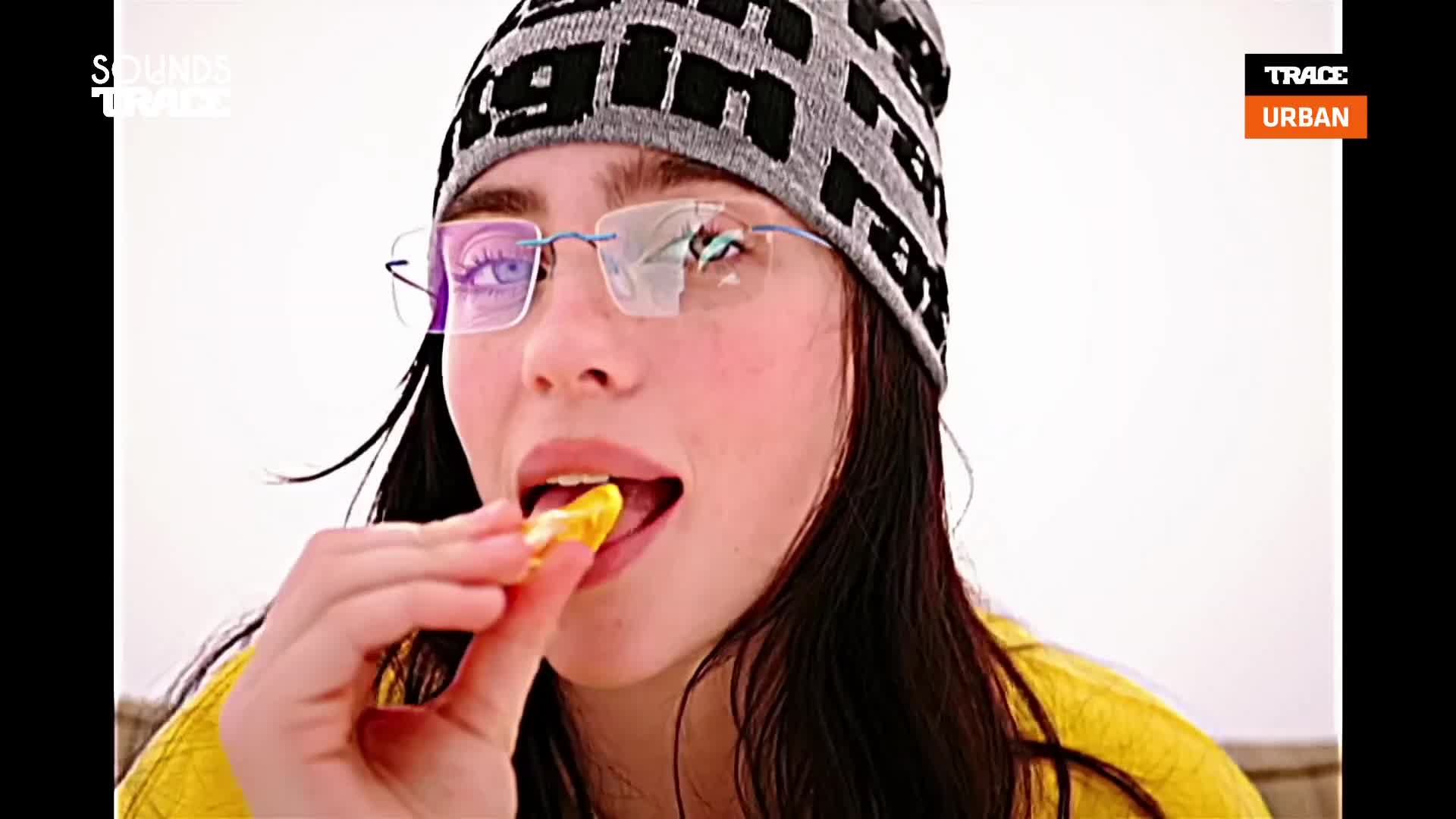 A young woman, wearing a beanie and glasses, bites into a yellow snack. The Trace Urban logo is visible in the corner.

