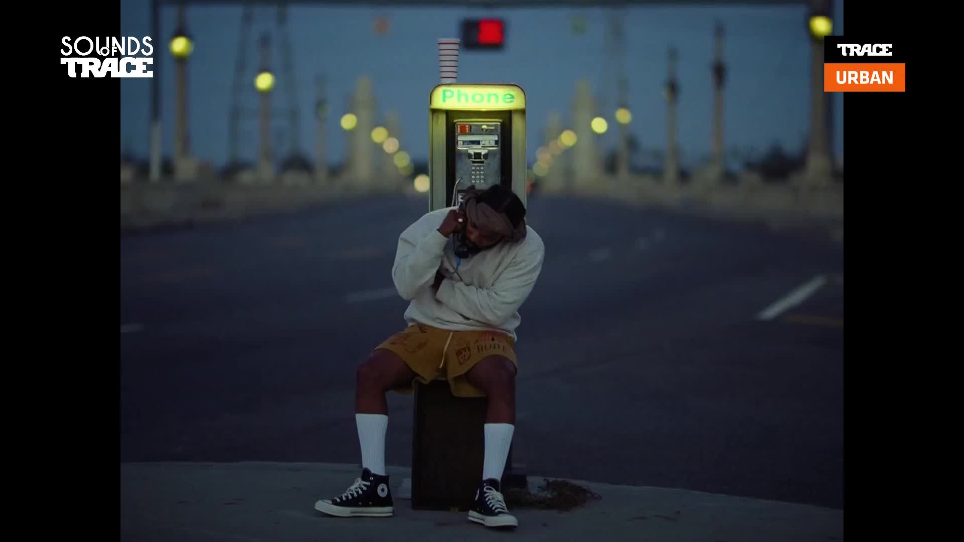 A man sits hunched on a phone booth pedestal, the receiver pressed to his ear. The streetlights glow behind him, casting a soft light over the empty bridge.
