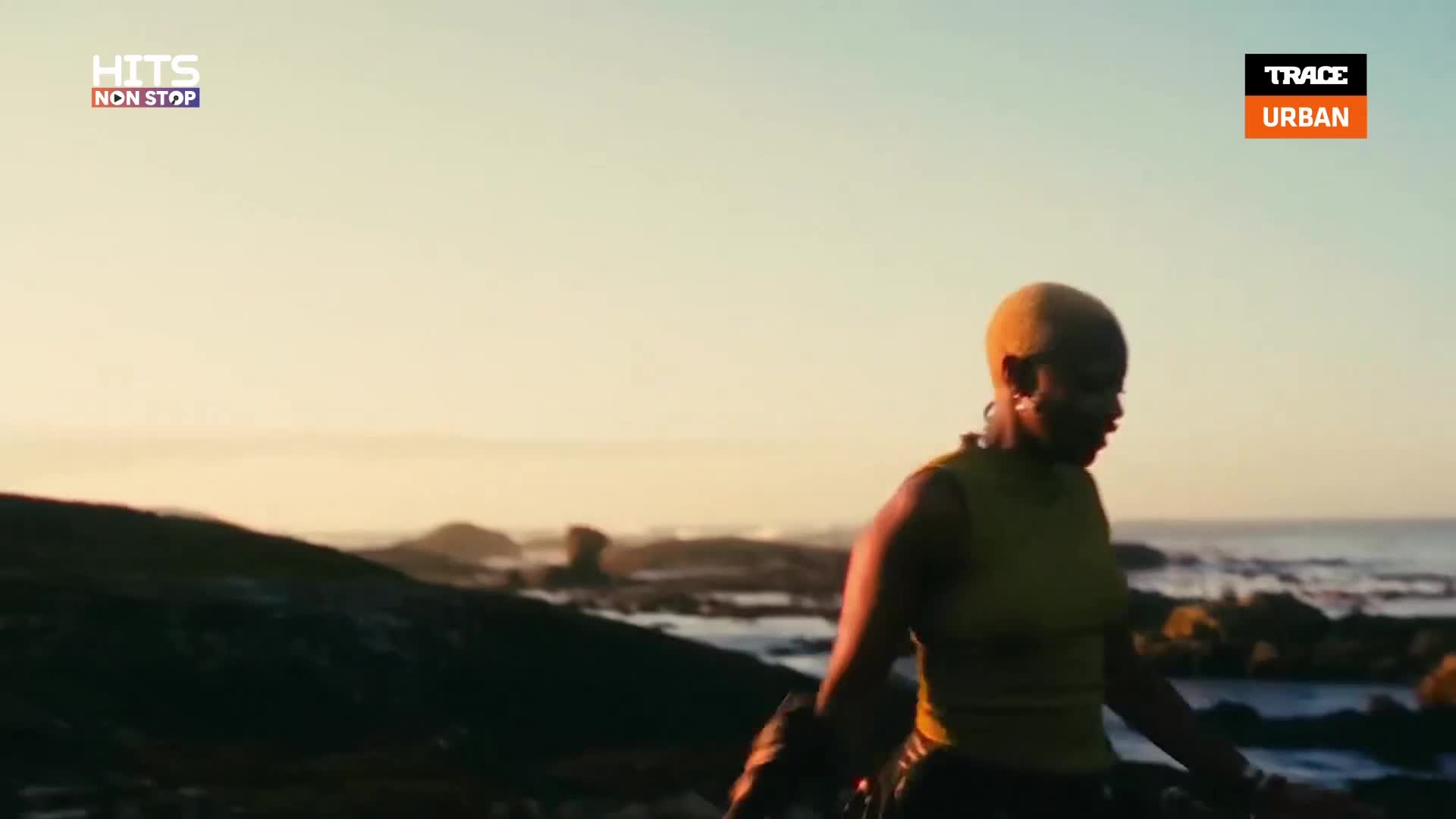 A woman with short, yellow hair walks along a rocky shoreline, the ocean stretching out behind her. The warm light of the setting sun bathes the scene, as if it's a music video on Trace Urban.
