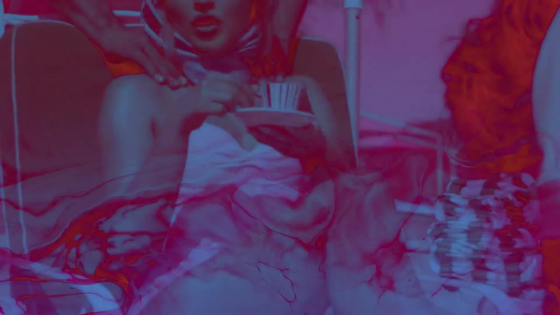 A woman reclines, holding a teacup, while someone massages her shoulders. The scene is bathed in a vibrant, stylized color palette, reminiscent of a music video.

