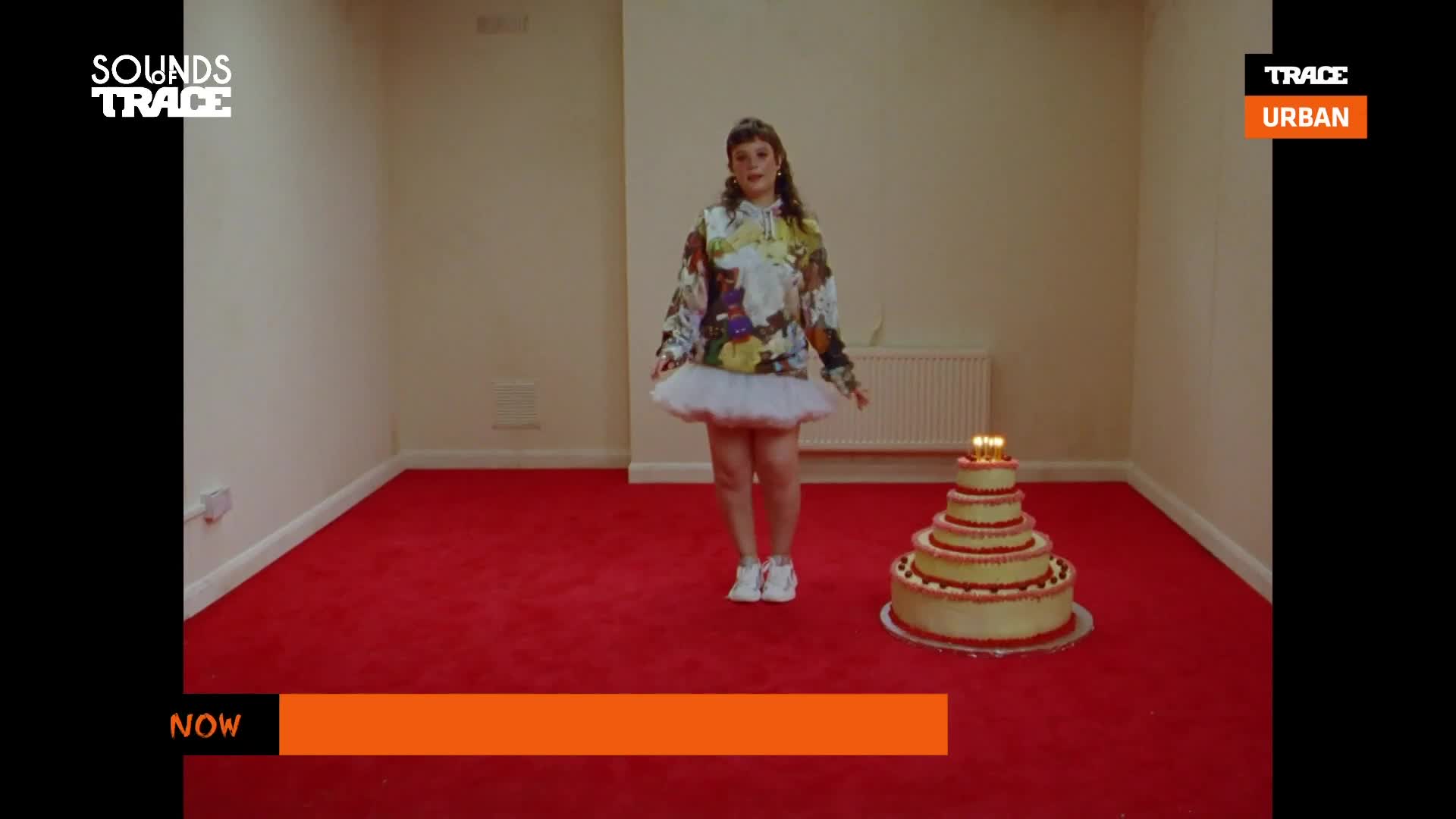 A young woman in a patterned hoodie and tutu stands in a stark room, facing a multi-tiered cake with lit candles. The Trace Urban logo is visible, and the scene looks like a music video.
