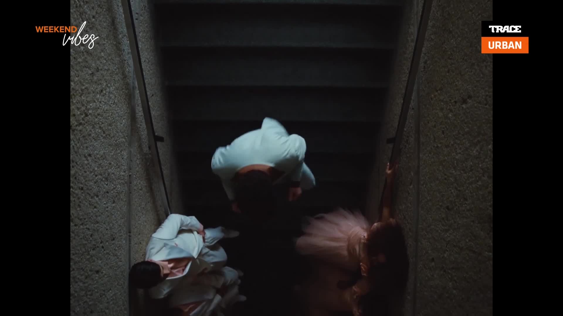 Three figures in white outfits tumble down a dark staircase, while a dancer in a pink tutu reaches out from the side. The scene is from a music video on Trace Urban, with "Weekend Vibes" branding.
