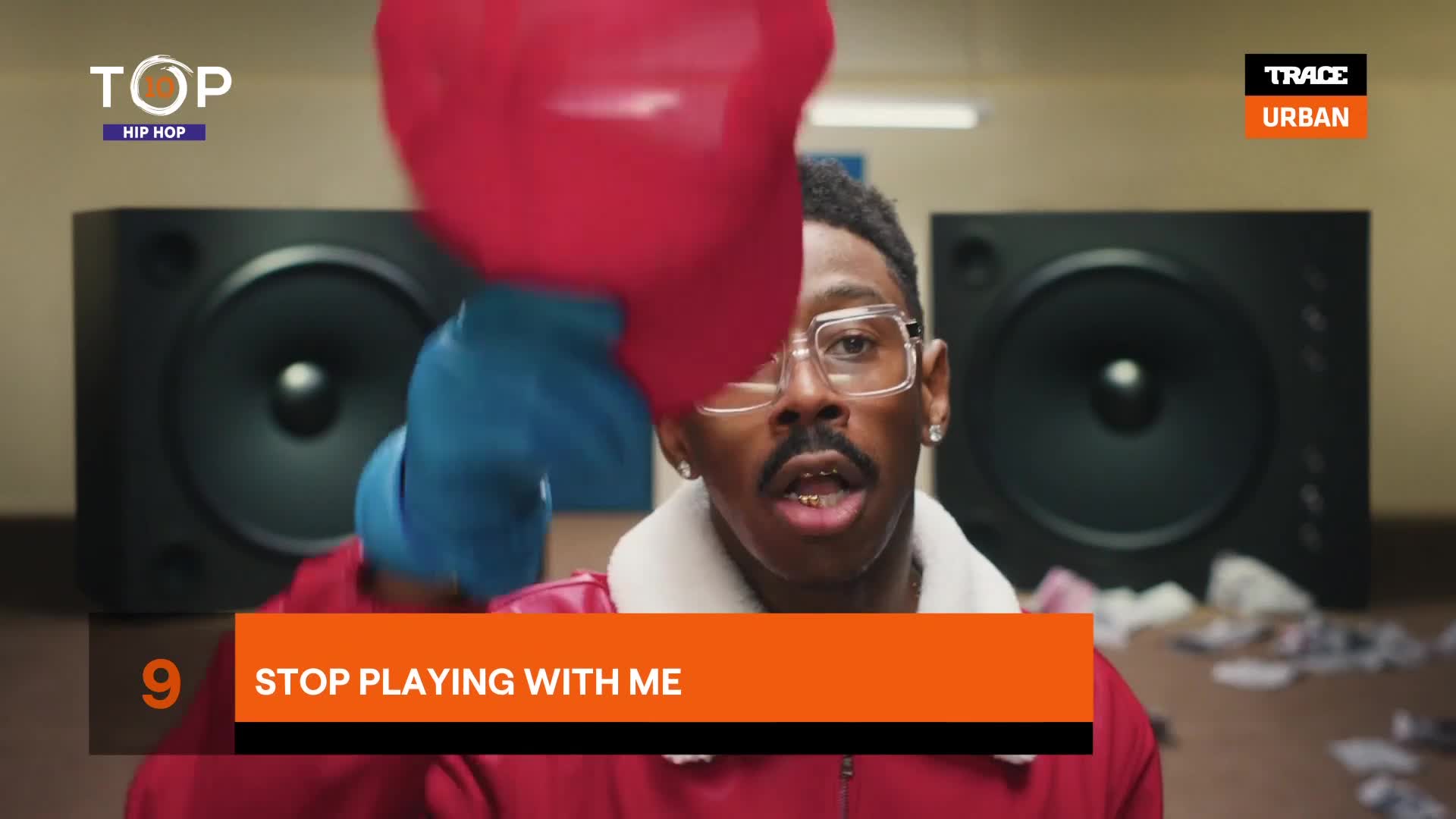 The man in the red jacket throws a red object, while wearing blue gloves, toward the camera. He's on the set of a music video, with a large speaker behind him.
