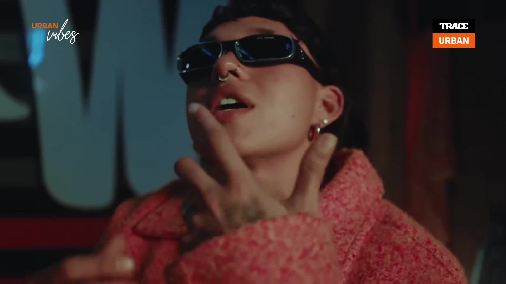 The person in the pink coat gestures with their hands, a silver ring glinting. Sunglasses cover their eyes as they look up, speaking into the camera.
