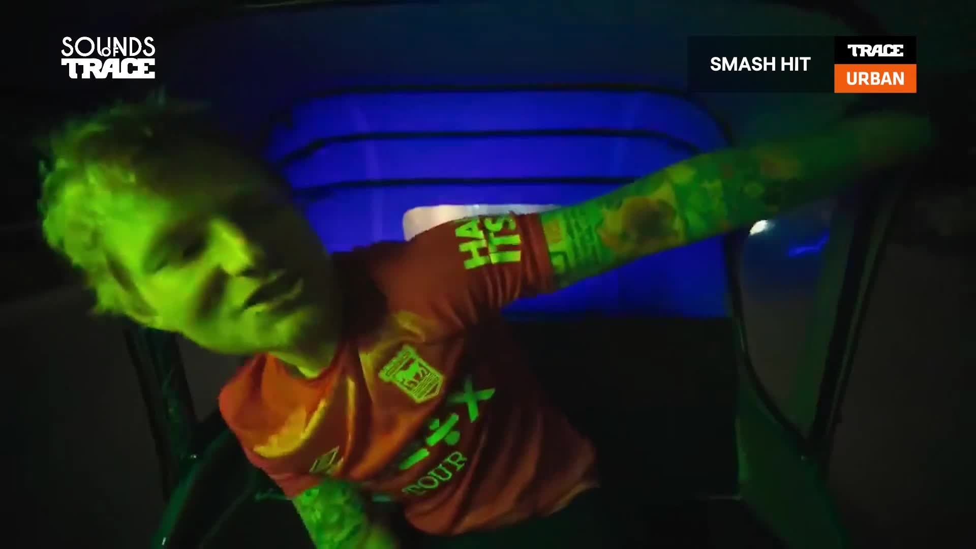 The man, illuminated in green, leans forward in a vehicle, his tattooed arm outstretched. A "Smash Hit" graphic and the Trace Urban logo are visible in the corner of the frame.
