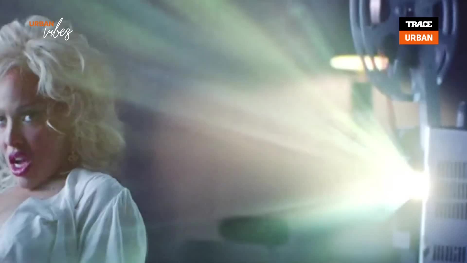 A bright beam of light shoots from a projector, illuminating the face of a woman with blonde hair. The Trace Urban logo is visible in the corner.
