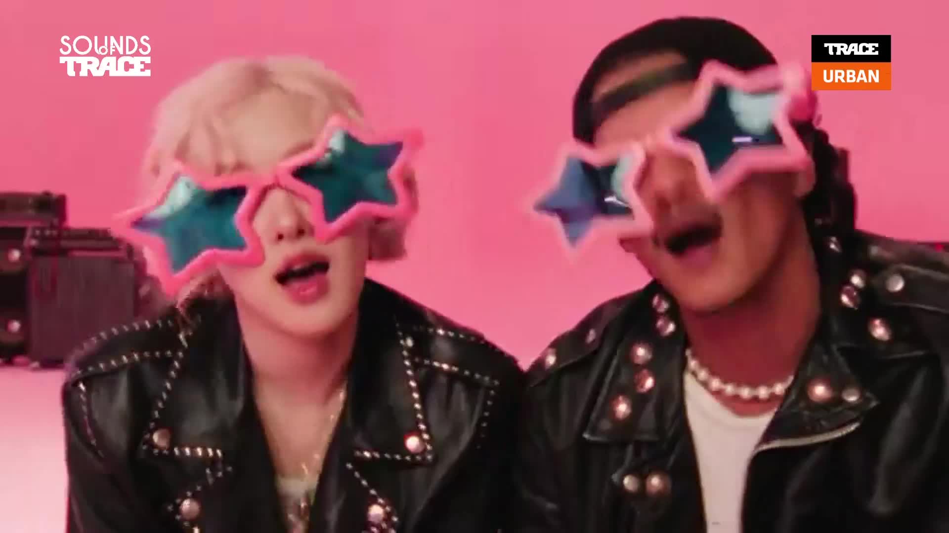 Two people, both wearing star-shaped sunglasses, sing into the camera, their leather jackets and the pink background adding to the playful vibe. The Trace Urban logo is visible, suggesting this is a music video.
