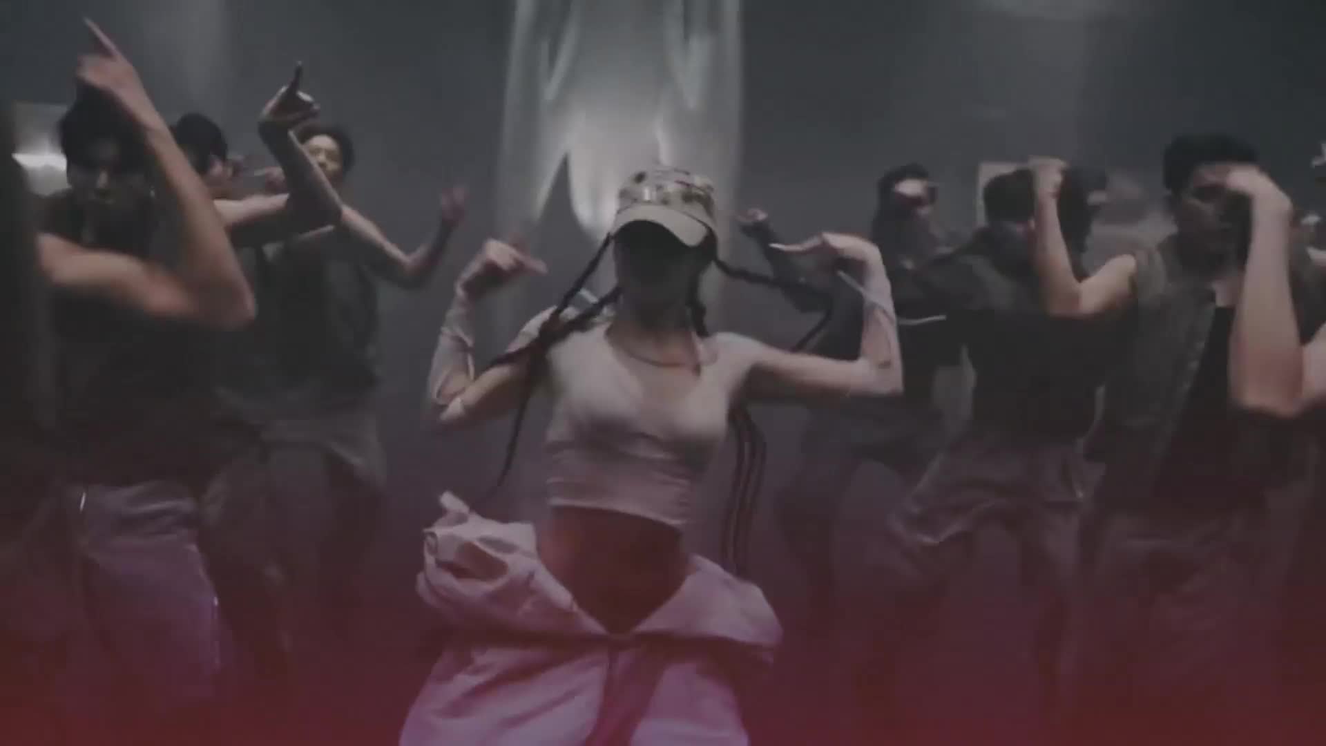 A woman in a cap and crop top is front and center, her arms raised as she dances. Behind her, a group of dancers mirrors her moves in a dimly lit space.
