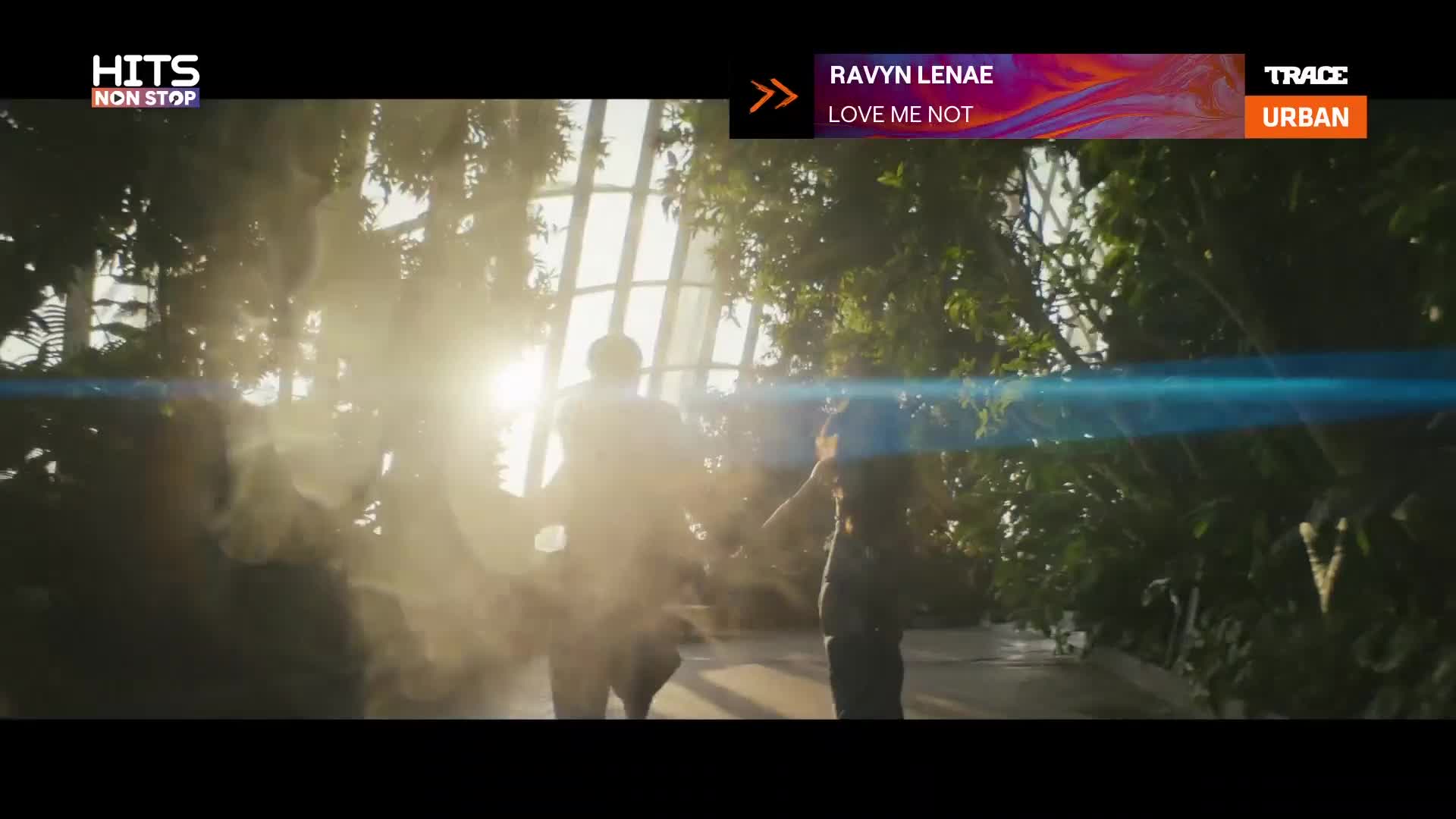 Two figures, backlit by bright sunlight, move through a misty, green space. The scene appears to be from a music video, playing on Trace Urban in France, for Ravyn Lenae's "Love Me Not."
