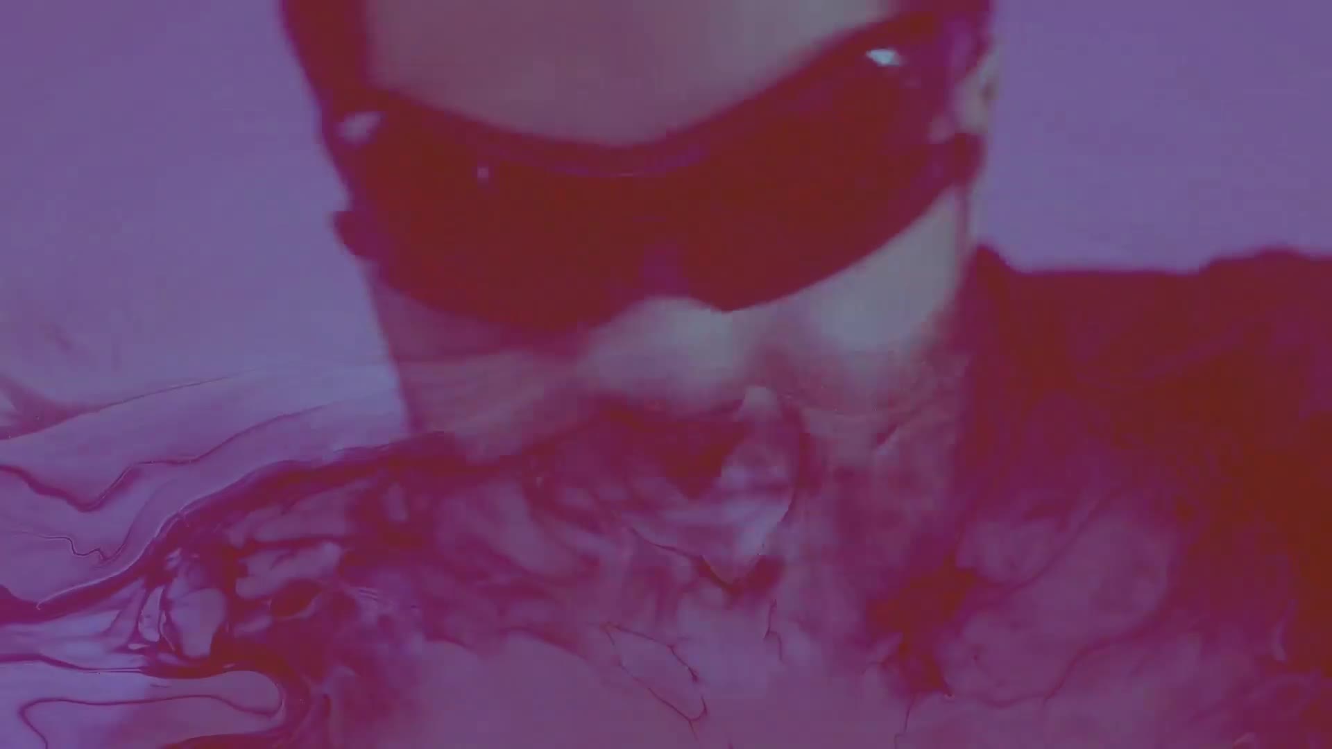 Dark sunglasses obscure the upper face, while swirling, reddish-purple liquid undulates below. The scene is bathed in a deep, moody light, hinting at a music video aesthetic.
