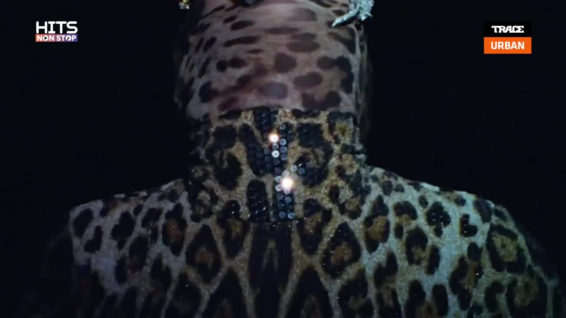 A leopard-print turtleneck and headscarf dominate the frame, with a vertical line of sparkling embellishments at the collar. The Trace Urban logo is visible, suggesting a music video or performance from France.
