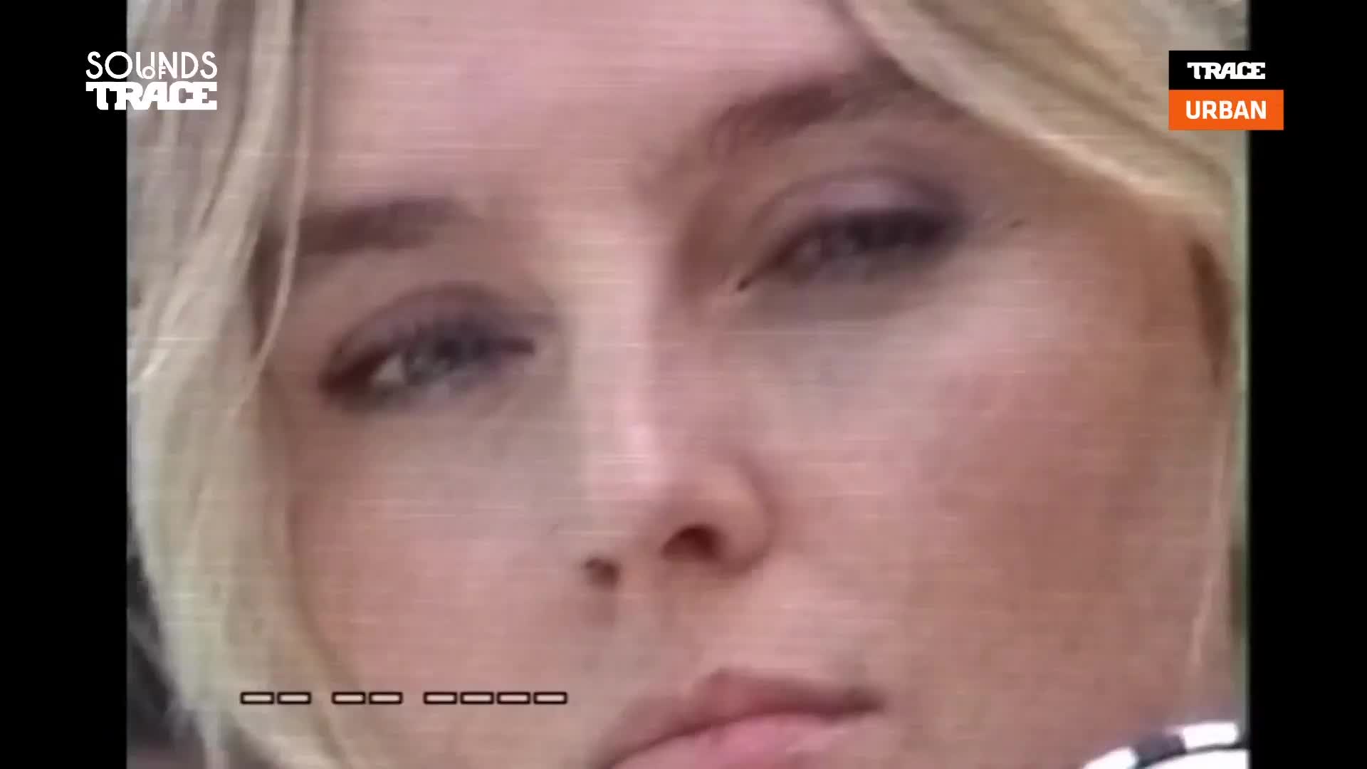 A woman with blonde hair looks directly at the camera, her expression subtle. The Trace Urban logo is visible in the corner, and a series of black boxes appear at the bottom of the screen.
