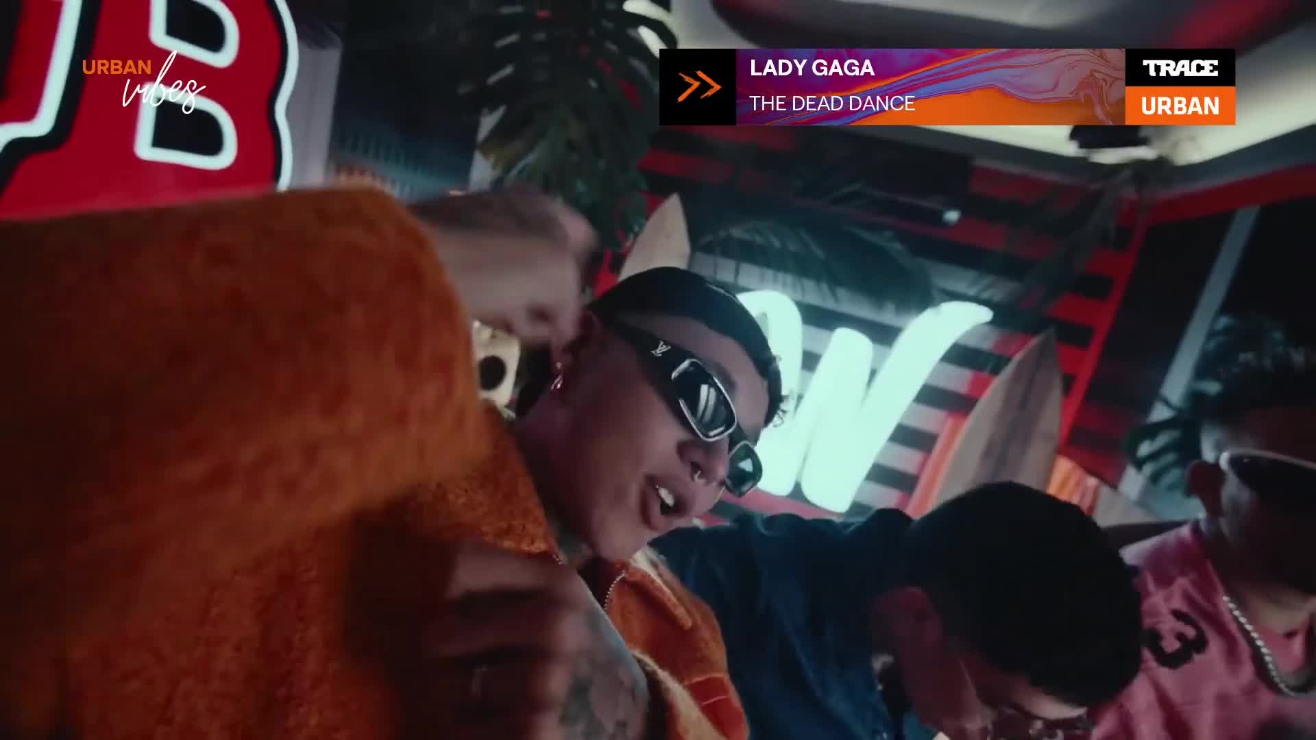 A young person, wearing sunglasses, sings into a microphone, their face illuminated by neon lights. The "Urban Vibes" logo and the Trace Urban logo are visible, suggesting a music video is playing.
