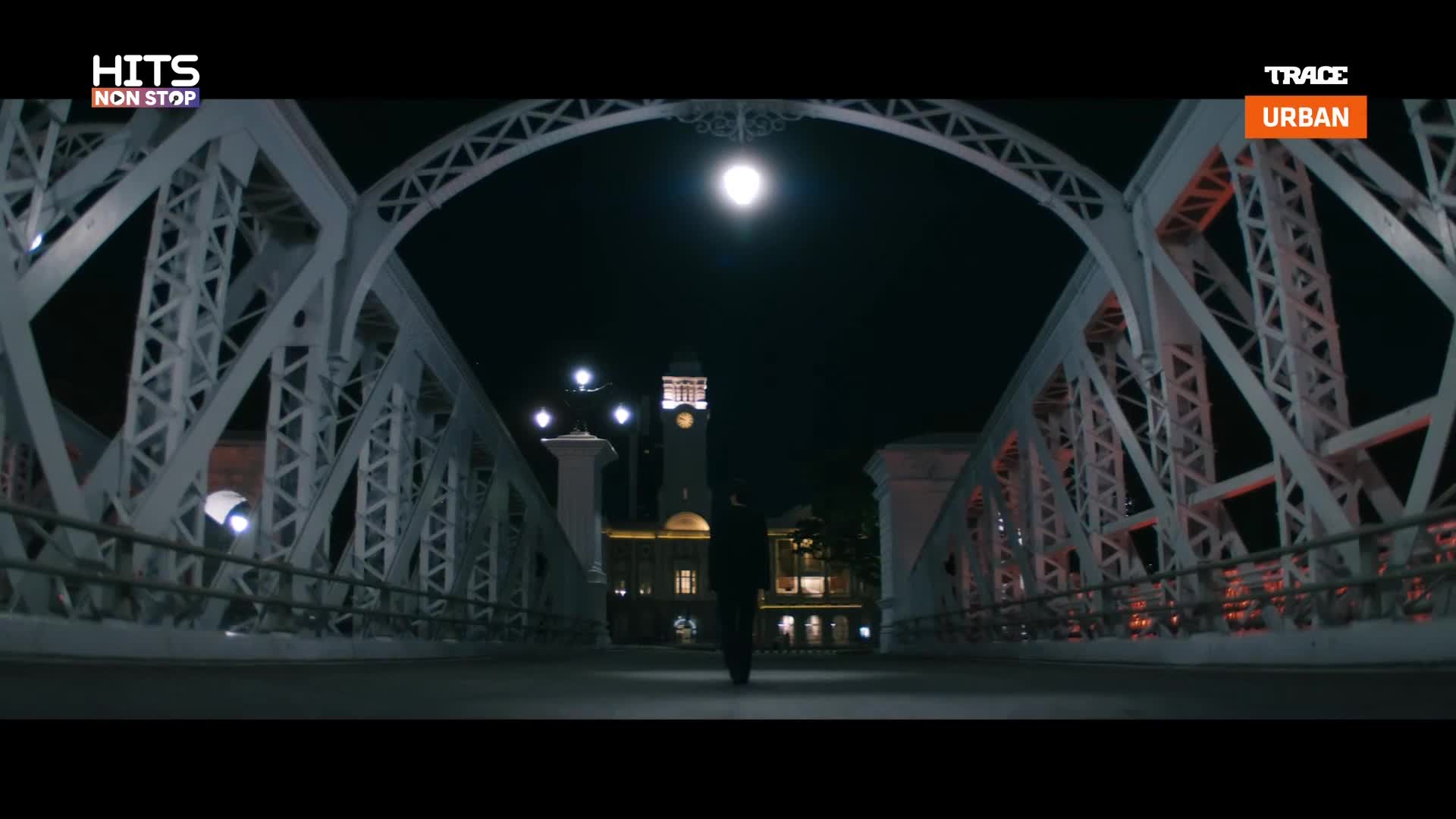 A lone figure walks toward the camera, silhouetted against the bright lights of a building in the distance. Overhead, the bridge's intricate metalwork frames the night sky and a full moon.
