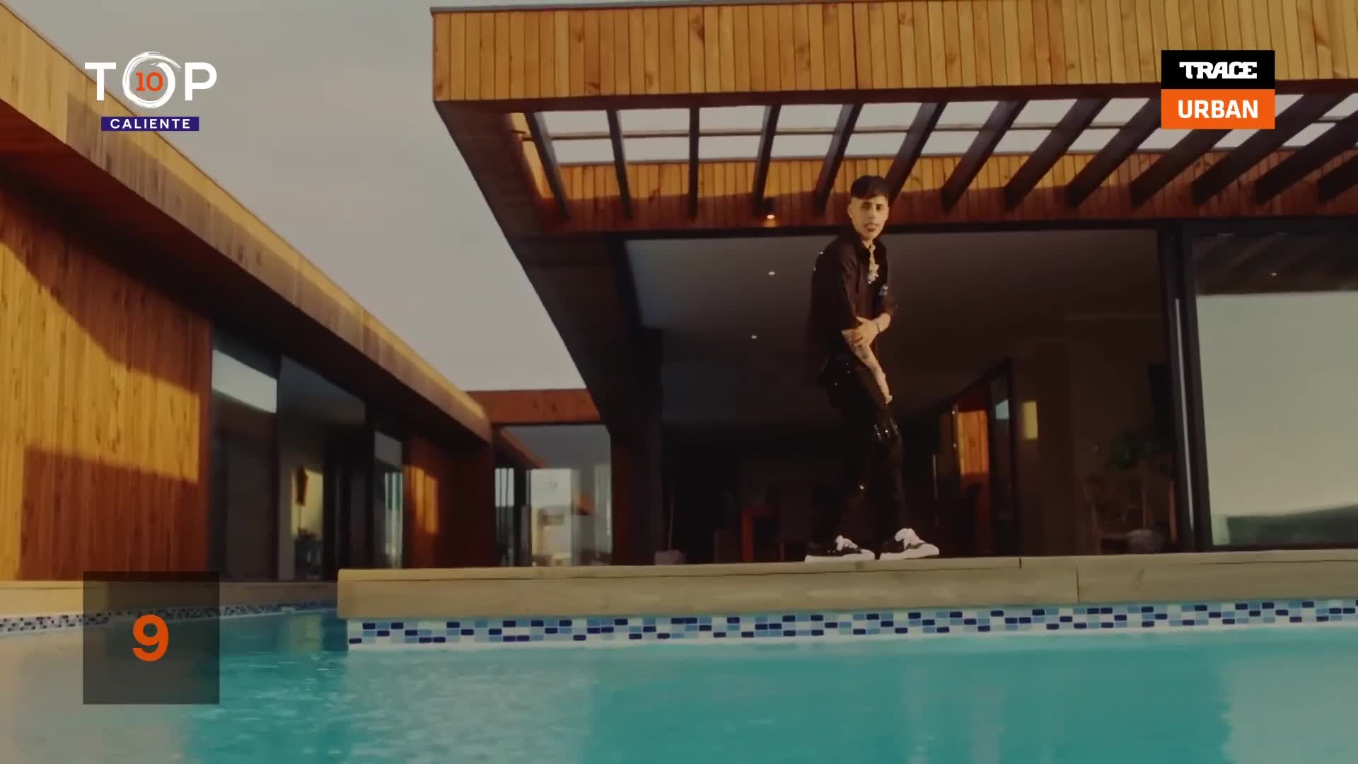 A young man in dark clothes is dancing on the edge of a pool, his moves framed by a modern house. The scene is playing on Trace Urban's Top 10 Caliente.
