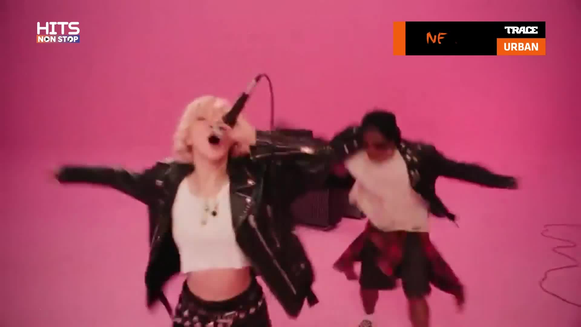 A woman with blonde hair belts into a microphone, arms outstretched in a leather jacket. Beside her, a person in a white t-shirt and a red plaid shirt around their waist moves energetically.
