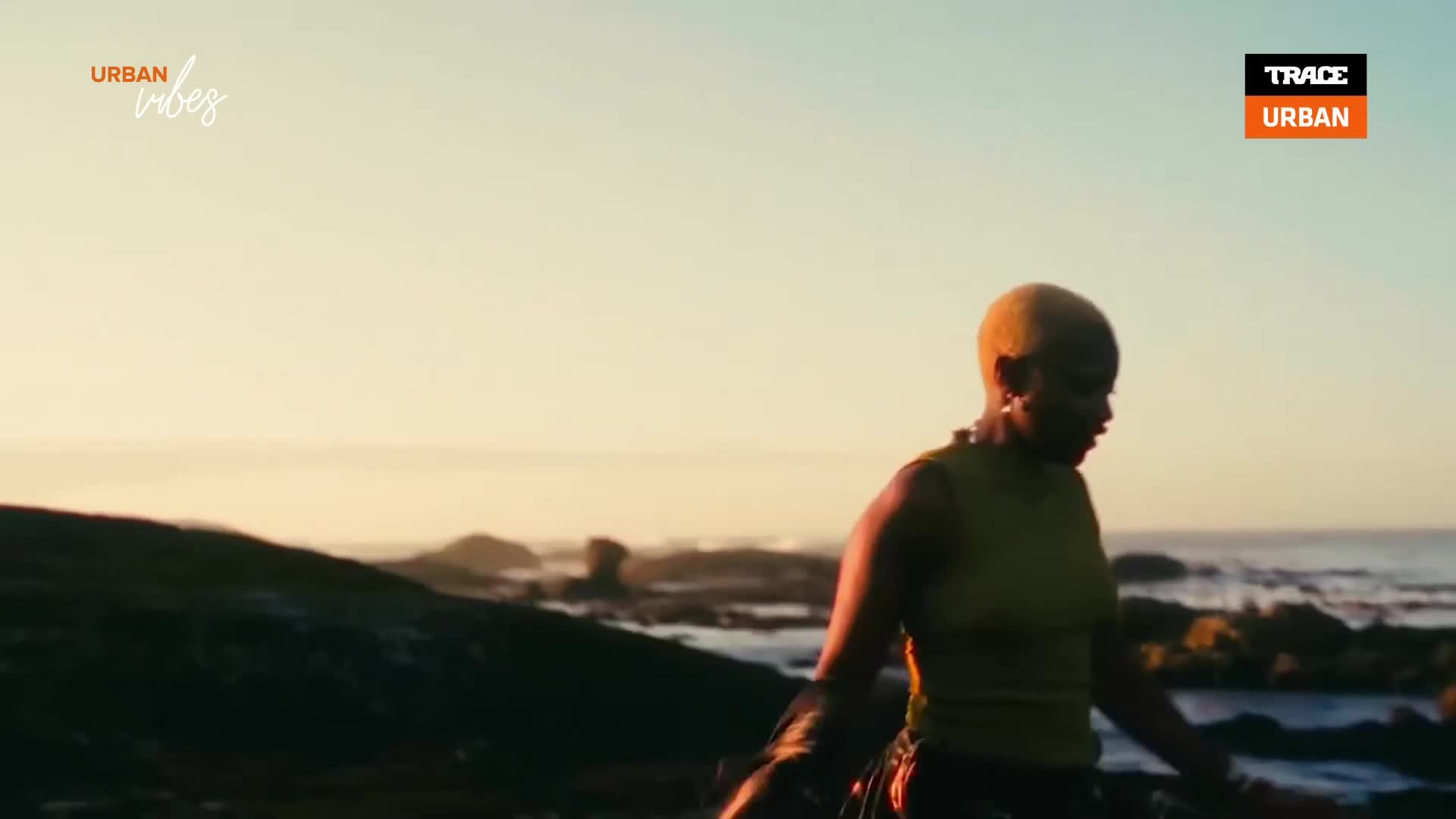 A woman with short, dyed hair walks along a rocky coastline, the sea stretching out behind her. The warm light of the setting sun bathes the scene, as seen on Trace Urban.
