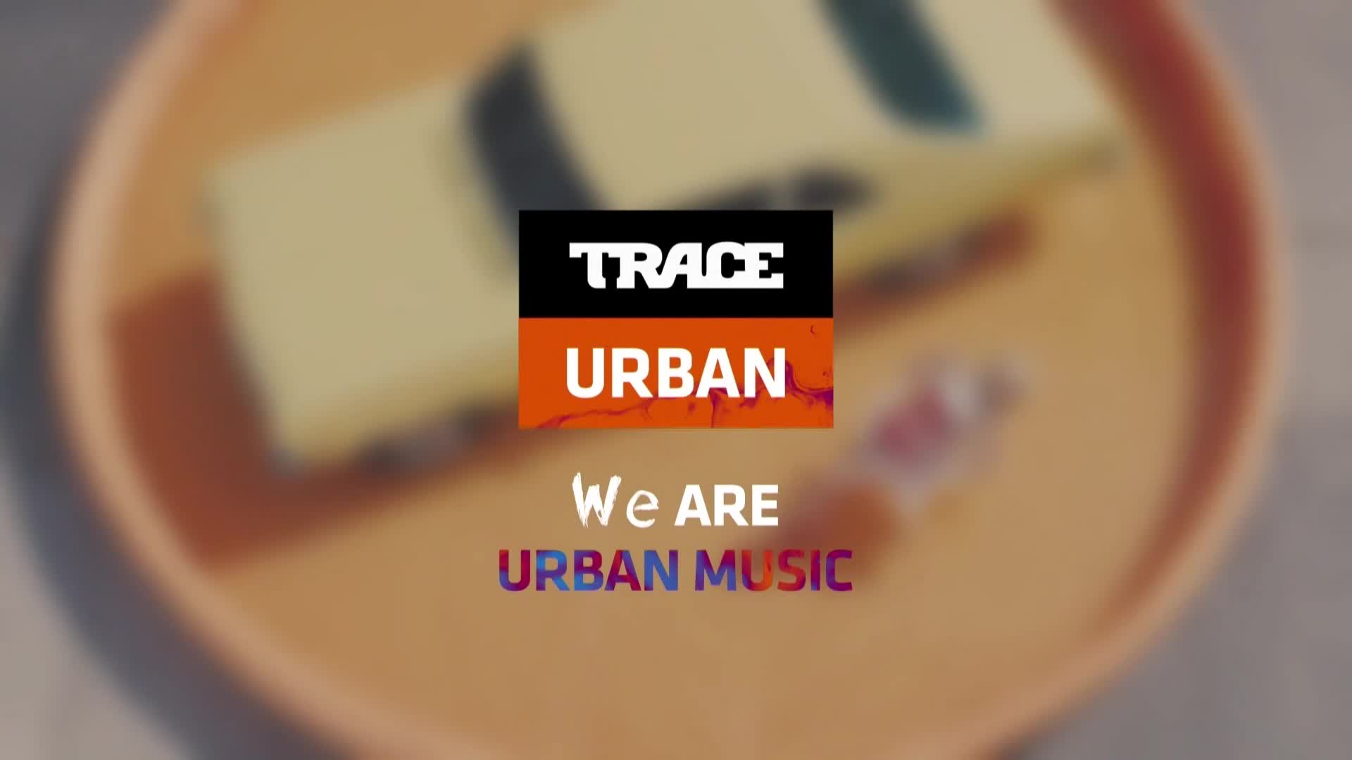 The Trace Urban logo, a black and orange rectangle, sits centered, with the words "We ARE URBAN MUSIC" below. A blurred, light-yellow rectangle and a small, translucent object are in the background.
