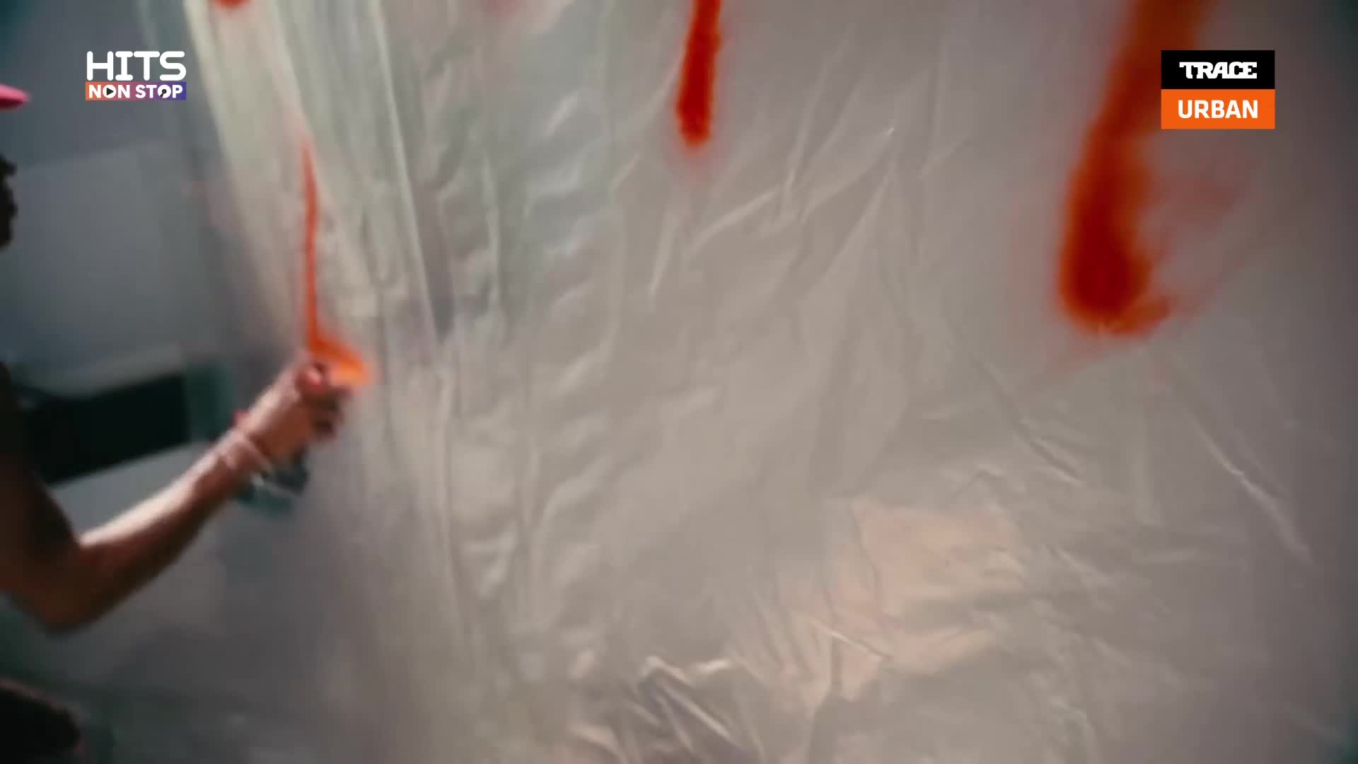 A person is spraying orange paint onto a sheet of plastic, creating long, dripping streaks. The scene is likely part of a music video or show, as indicated by the "Trace Urban" logo.
