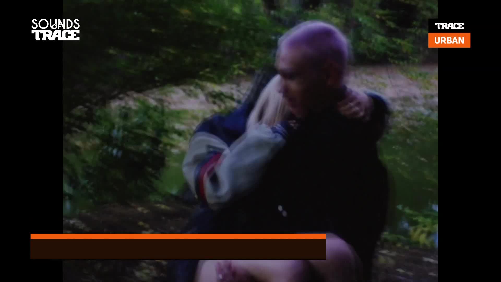 A person with purple hair holds someone close, their arms wrapped around the other's shoulders. The scene is set in a blurry, green-tinged outdoor space, possibly a music video on Trace Urban.
