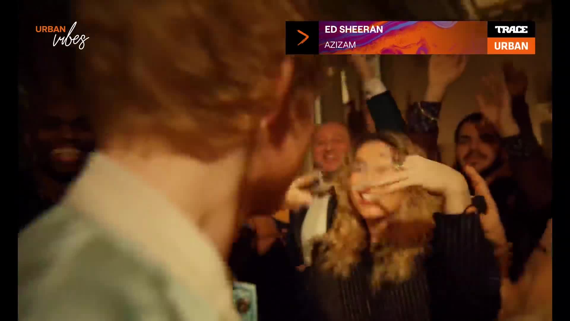 A crowd of people is celebrating, their arms raised in the air. The screen shows "Azizam" by Ed Sheeran on Trace Urban.
